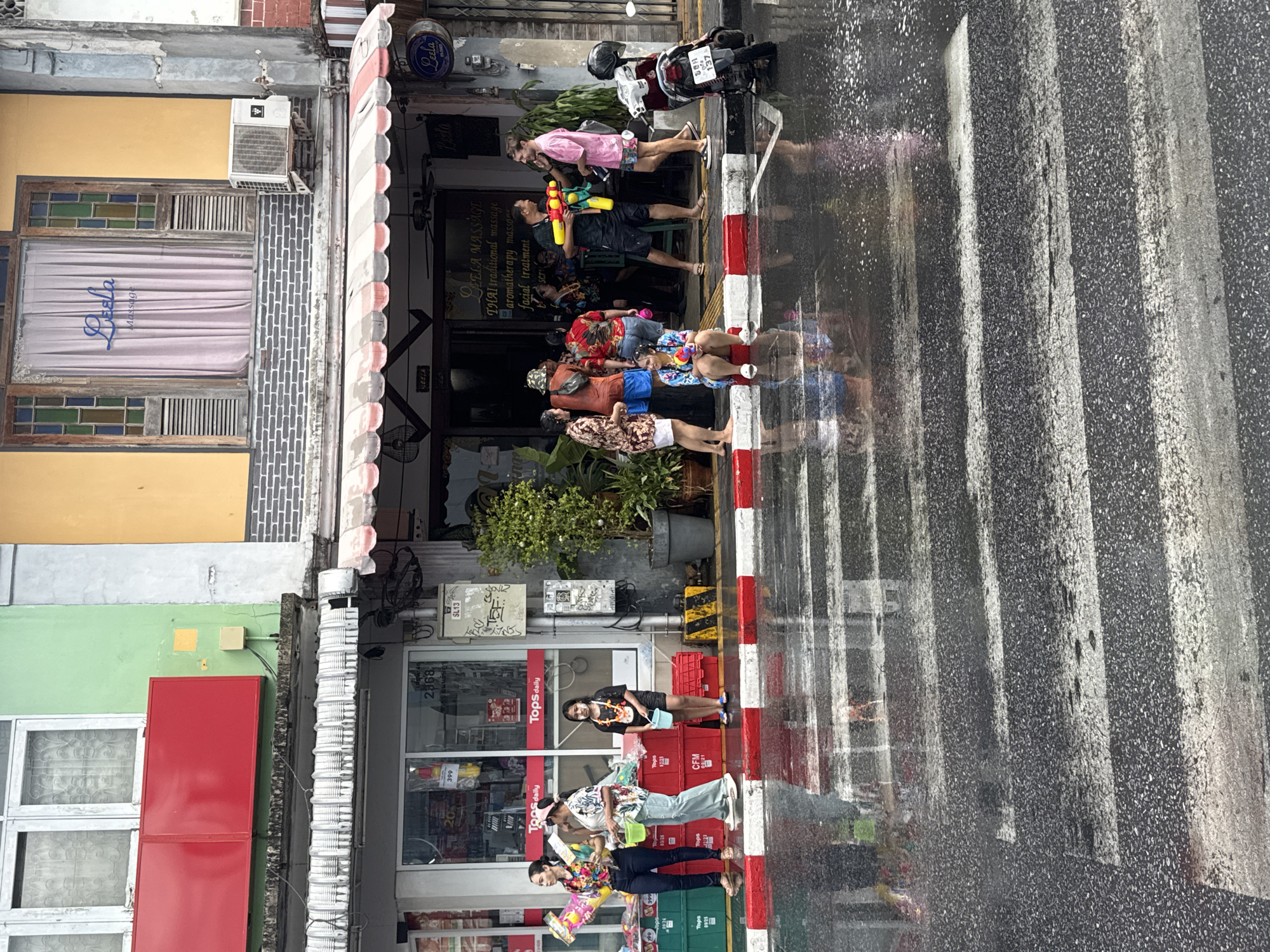 Here is a description of the image:
This scene is a street view in Phuket City Municipality, Thailand, likely captured during a festival. A group of people stands on the sidewalk and edge of the road in front of commercial establishments, including a "Tops daily" store and a business identified as "LEELA MASSAGE". Several individuals are seen holding water guns, indicating participation in a water-related event, possibly the Songkran festival. The street is wet, and the crosswalk is clearly visible on the road. A motorcycle is parked near the "LEELA MASSAGE" building. The buildings have a variety of architectural elements, including awnings, windows, and different colored facades.