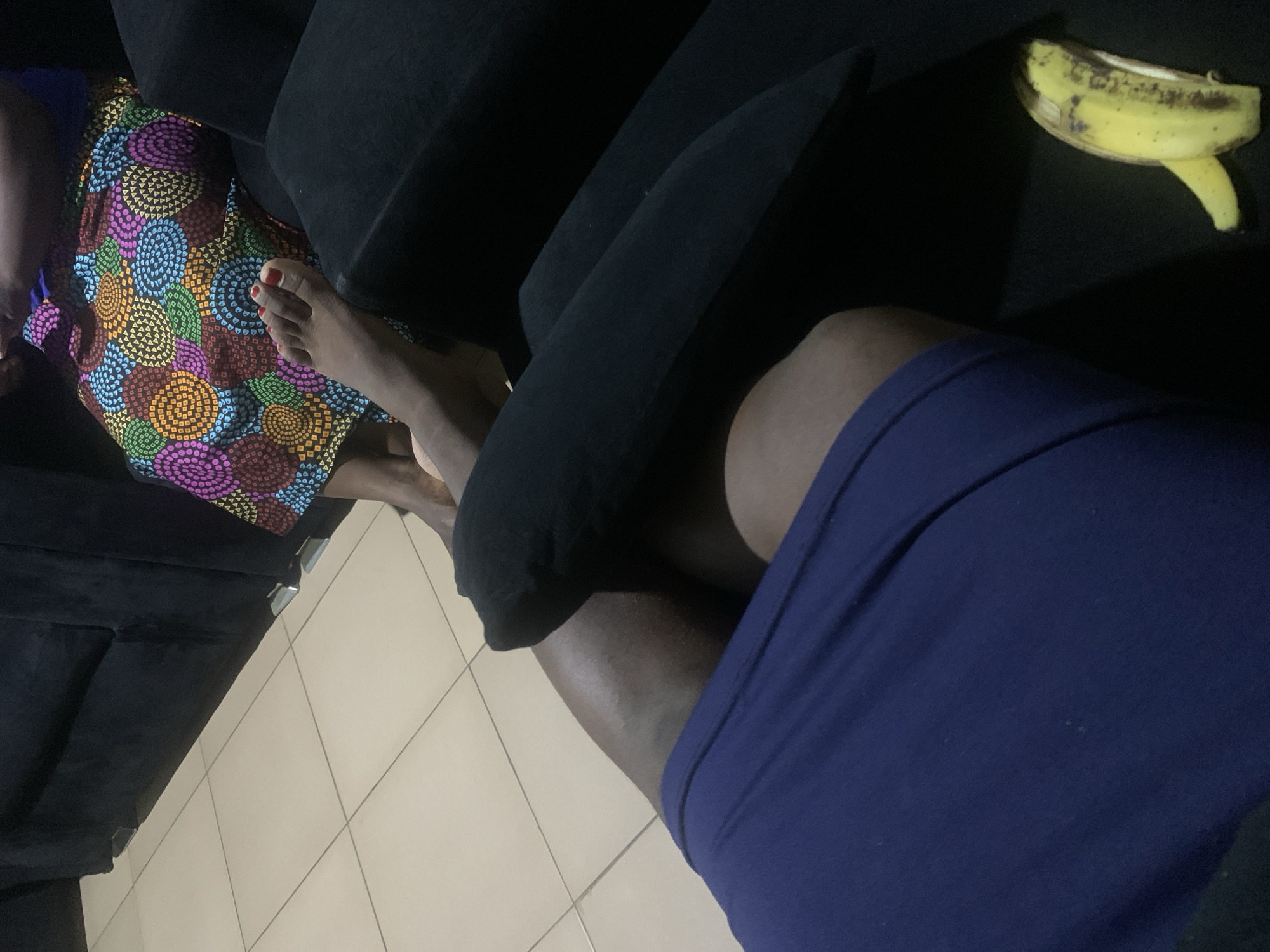 The image depicts a close-up view of a person's legs and feet, positioned on a dark couch. To the left, a person's bare legs are visible, wearing a dark blue garment. To the right, another person's feet are visible, wearing red nail polish and resting against a colorful, patterned skirt. A peeled banana lies on the couch to the right of the legs. The floor appears to be tiled. The setting is indoors.