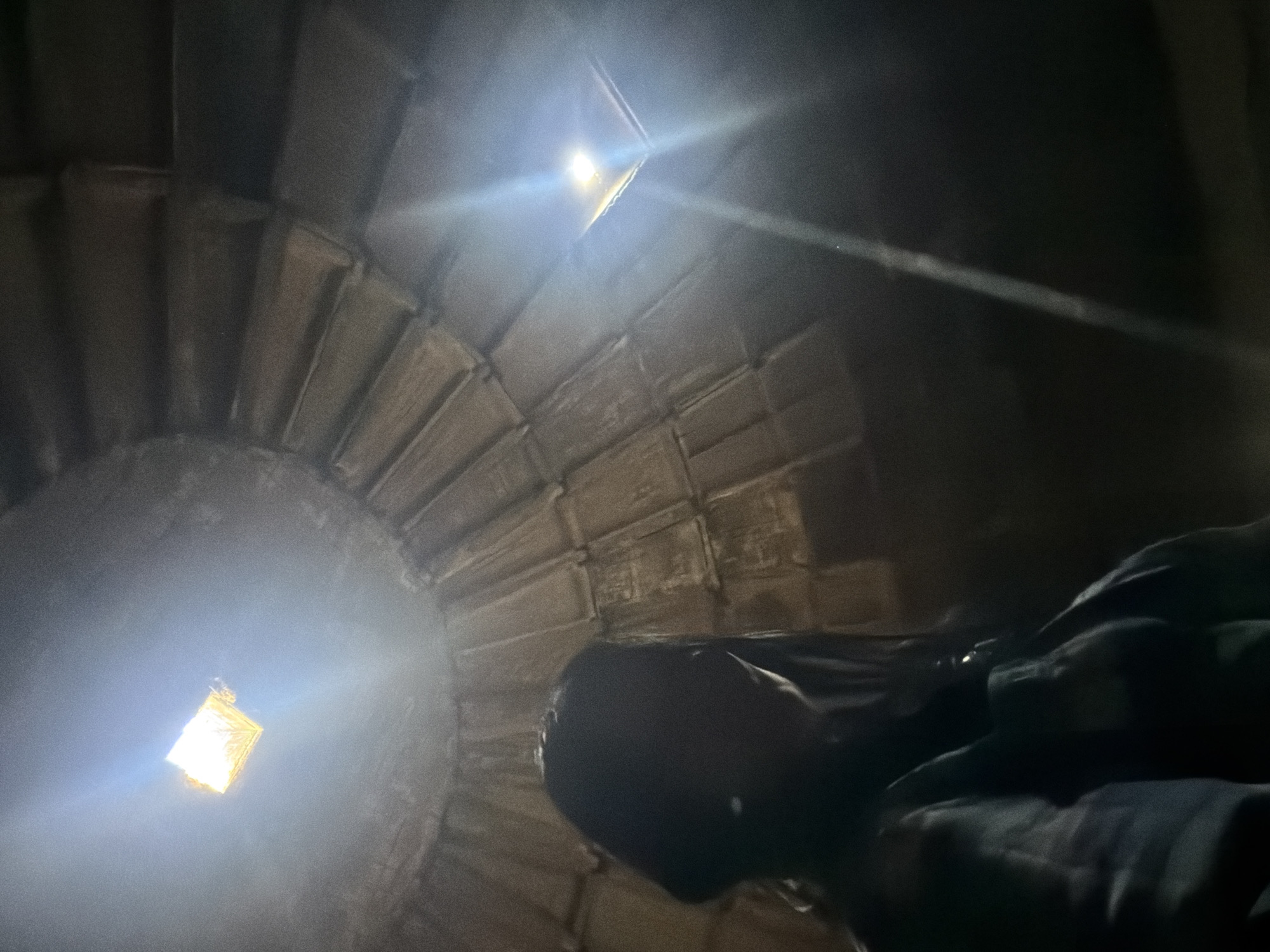 The image appears to be taken from inside a tower or shaft, looking upwards towards a light source. The interior is constructed of layered stones, creating a circular or spiral structure. There is a bright square opening at the top, presumably letting in light. The image is dark overall, with the primary illumination coming from the opening above. A person's head is visible in the foreground. The specific location is not determinable from the image alone.