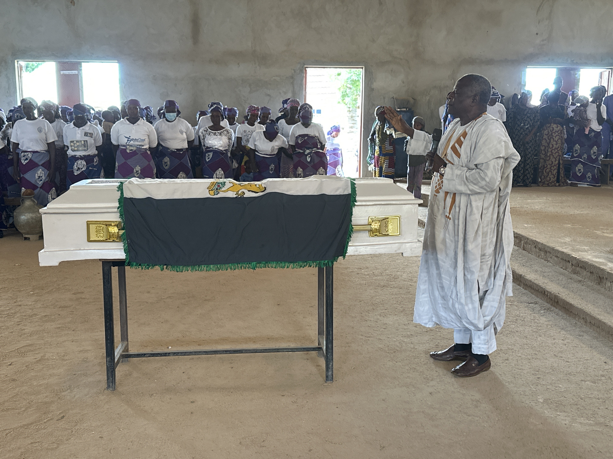 In Kufana, Nigeria, a somber gathering unfolds inside a simple, unpainted building. A white coffin, draped with a dark cloth trimmed in green fringe and featuring a green and yellow emblem, rests on a black metal stand in the foreground. A man in a light-colored traditional robe and pants, his hands raised in a gesture of prayer or blessing, stands to the right of the coffin, facing a congregation of people.
The congregation, primarily women dressed in white tops and vibrant purple and grey patterned skirts and headscarves, stands behind the coffin. Some individuals wear face masks. The background reveals open doorways and windows, hinting at the time of day. The overall atmosphere appears to be one of solemnity and reverence, likely a funeral or memorial service. The ground is a sandy, earth-toned surface. Visible text includes "WhiteCloud" on one of the woman's shirts.