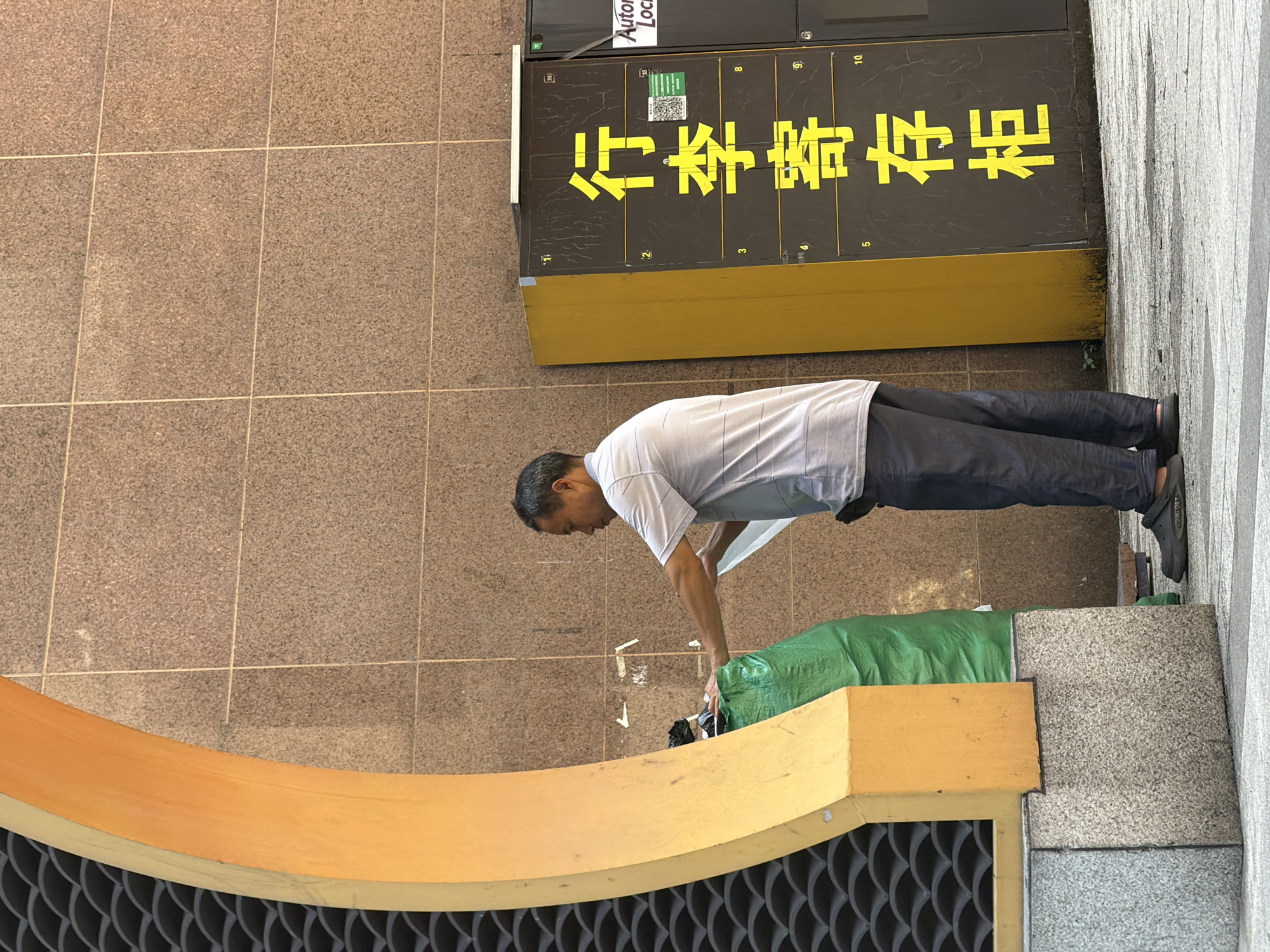 A man stands outdoors in front of a tiled wall, a yellow-painted structure with a curved opening, and a yellow locker unit with Chinese characters. The man, wearing a light-colored short-sleeved shirt and dark pants, is reaching into a large green trash bag with his right hand while holding a white document or paper in his left hand. The locker unit is labeled with "行李寄存柜" in large yellow characters, indicating it is a luggage storage locker. It also displays numbers and a QR code. The ground is paved with concrete and stone tiles. The scene appears to be in Yuzhong District, China.