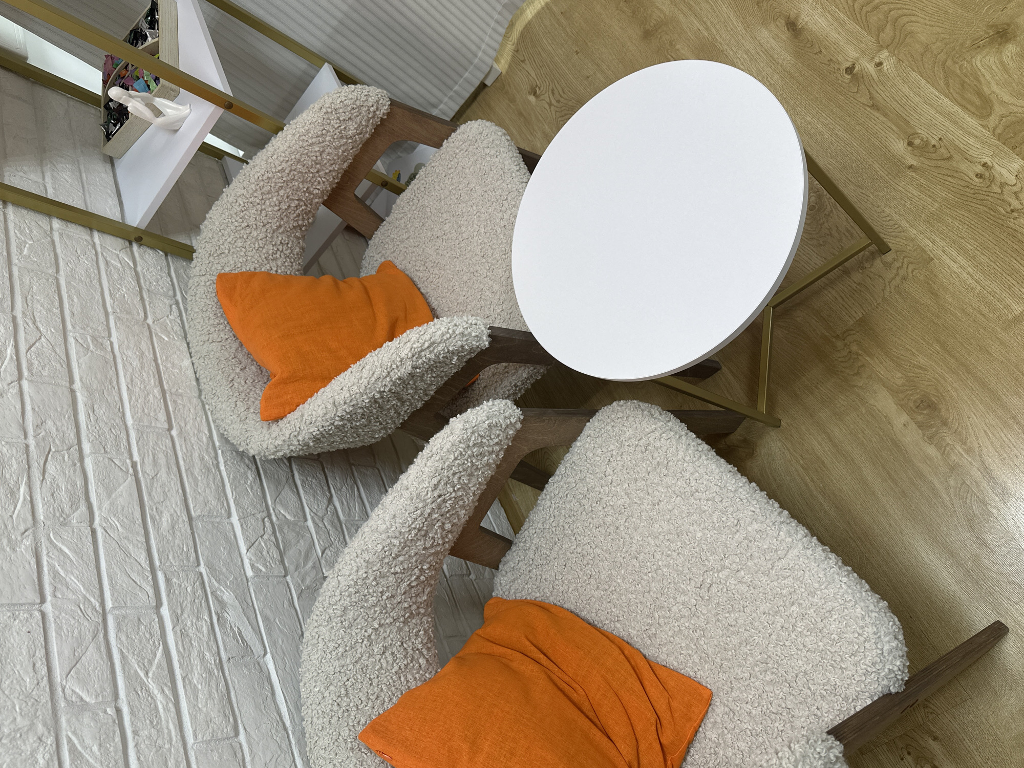 A top-down shot depicts two white, sherpa-like upholstered chairs with orange square pillows, positioned around a small, round, white table with gold-colored legs. The chairs are set against a white brick wall on the left and a light brown wooden floor on the right. A white shelf with gold trim is visible above one of the chairs on the left. The location is Chaikovsky, Russia.