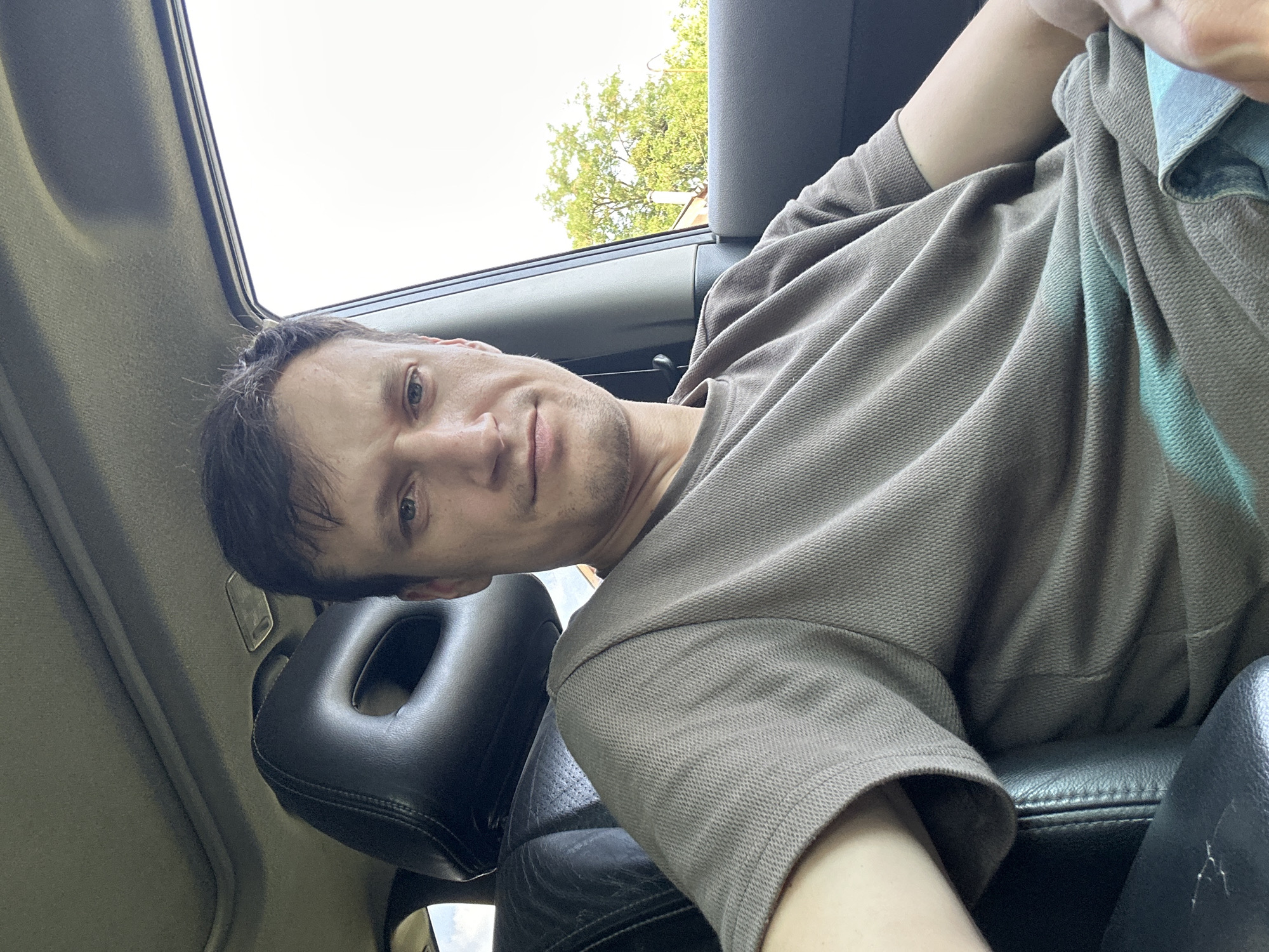 A medium shot selfie shows a man sitting in the driver's seat of a car. The man has short, dark brown hair, a fair complexion, and is looking directly at the camera with a neutral expression. He is wearing a short-sleeved, brown, textured t-shirt. The interior of the car is visible, including the black leather headrest of the driver's seat and a portion of the sunroof. The exterior of the car is partially visible through the passenger side window, showing green trees and a bright sky, indicative of an outdoor location. The lighting suggests it is daytime. The background detail of trees and what appears to be a building suggests this may be a residential area. Without further information, the specific location in Makiv, Ukraine, cannot be precisely determined beyond being within the city limits.