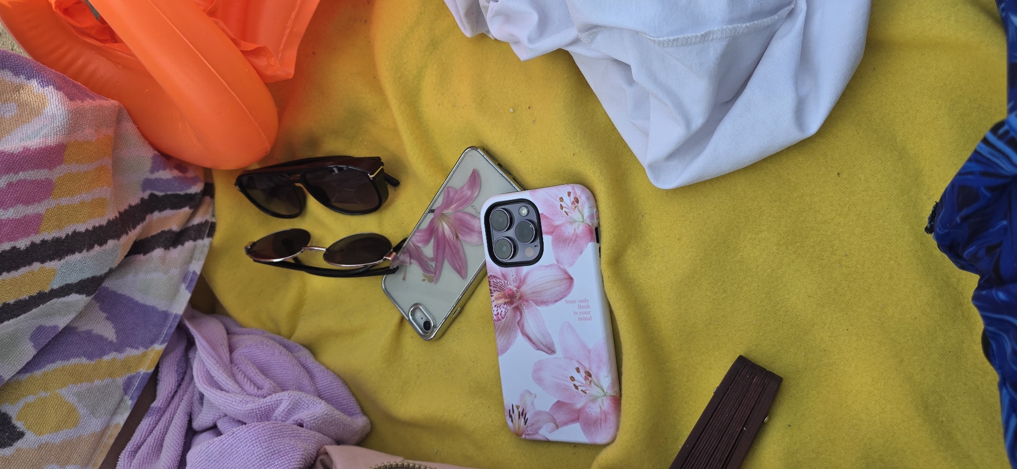 The image depicts a close-up, overhead view of various items laid out on a yellow blanket, likely in an outdoor setting such as a beach or park in Бактуу-Долоноту, Kyrgyzstan. The scene is cluttered with personal belongings. In the upper left, a folded towel with a multicolored striped pattern is visible, adjacent to a crumpled lavender towel. To the right of these, an orange inflatable ring is partially visible.
The primary focus of the image is on two cell phone cases and a pair of sunglasses. One phone case, lying flat, features a clear casing with pink floral imprints. A second phone case, placed slightly below and to the left, also displays a pink floral design, with a prominent camera bump. A pair of sunglasses rests on top of the clear phone case, with the lenses facing upwards.
Other objects present include a small, dark brown wooden fan lying on its side in the lower left quadrant of the image. A piece of white fabric is draped in the lower right corner. The overall impression is one of personal effects casually arranged on a bright yellow surface. The exact location within Бактуу-Долоноту is not discernible from the visual information alone.