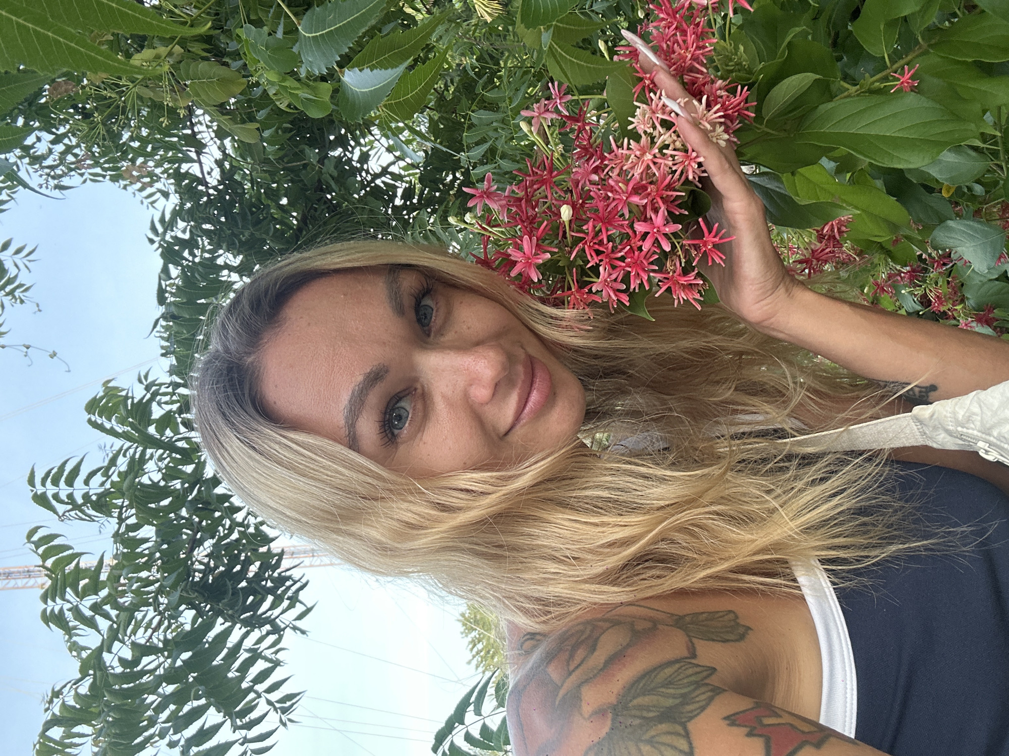 A blonde woman is holding a bunch of pink flowers in her right hand. She is wearing a navy blue and white tank top. She has tattoos on her left arm. The woman is in front of lush green foliage. The background shows a clear blue sky and what appears to be a cell tower. The image was taken outdoors in Phan Thiết, Vietnam.