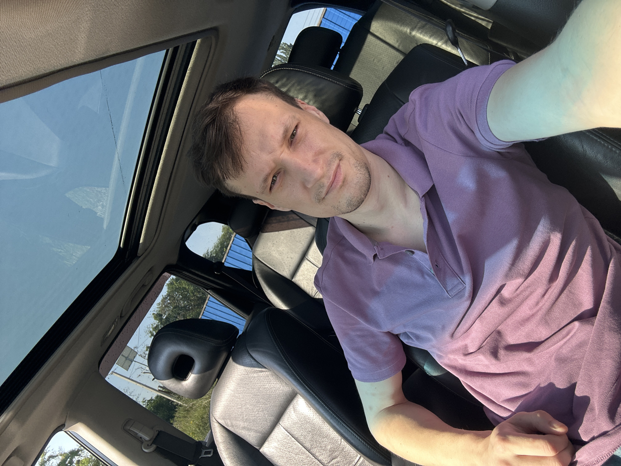 A man is sitting in the driver's seat of a car, looking at the camera. He has short brown hair and is wearing a purple polo shirt. The car has a sunroof that is open, and the sky is visible through it. The car's interior is dark, with black leather seats. Outside the car, trees and a blue fence can be seen. The image appears to be taken during the daytime, as evidenced by the bright sunlight. There is no indication of the specific location within Shatava, Ukraine.