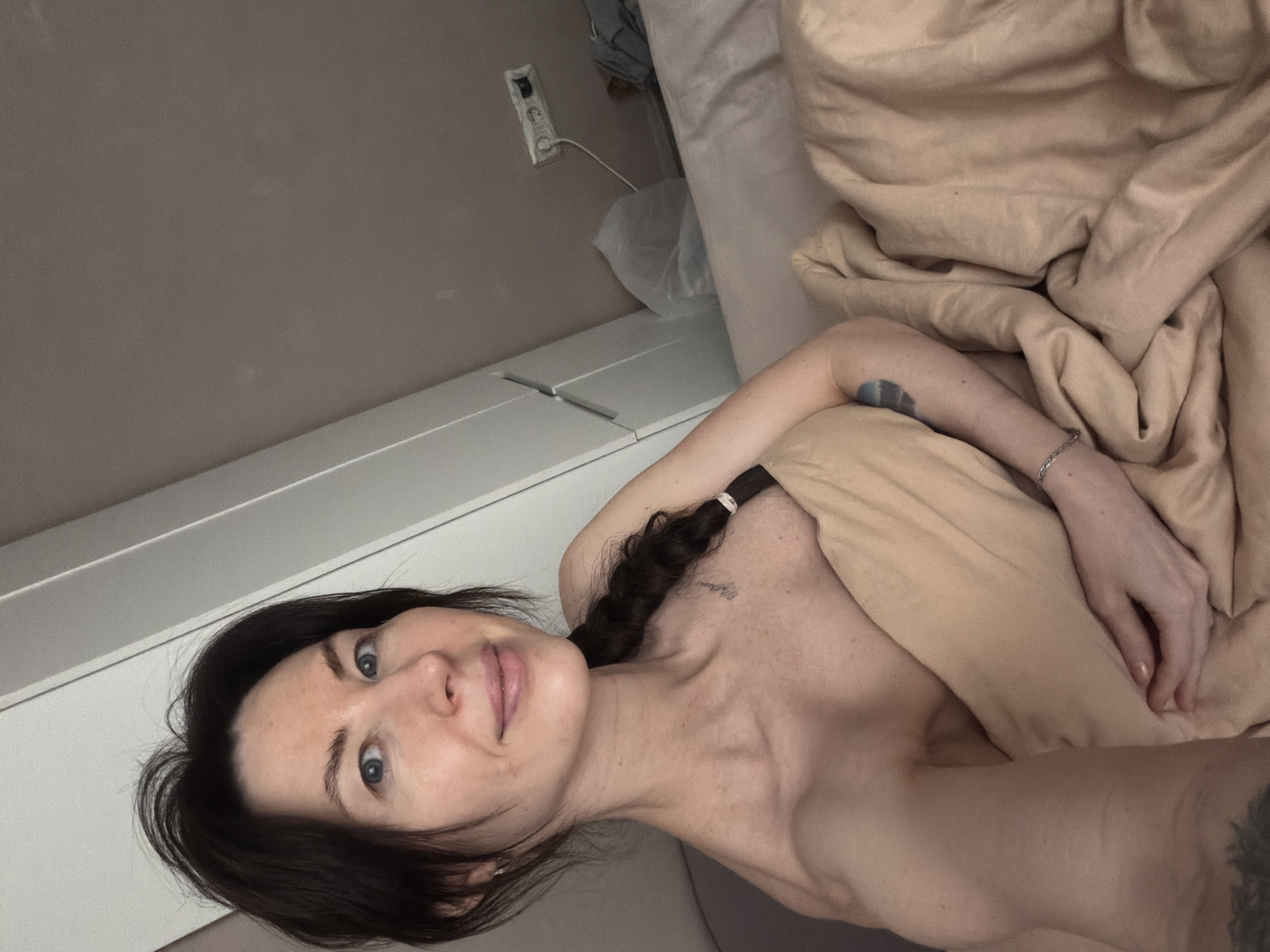 A woman with dark hair braided on one side lies in bed under a tan duvet. The background features a light-colored wall, a white cabinet, and an electrical outlet. No details indicate the image was taken in Perm, Russia.