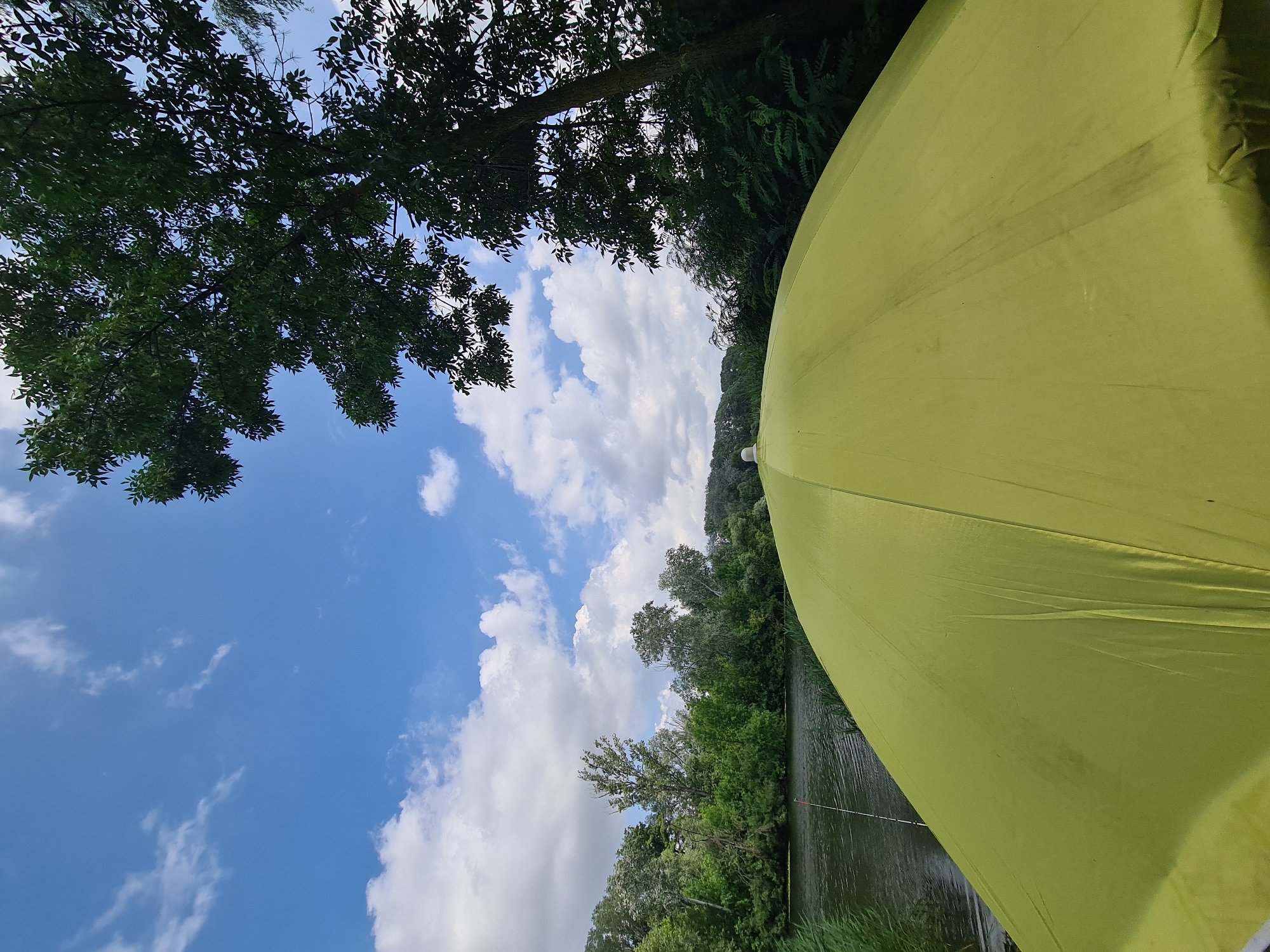 The image captures a serene waterside scene in Potoky, Ukraine, dominated by a vibrant lime-green umbrella in the foreground, its fabric stretched taut and curved upwards. Beyond the umbrella, a tranquil body of water reflects the bright blue sky dotted with fluffy white clouds. Along the opposite bank of the water, a dense, verdant forest stretches as far as the eye can see, creating a natural barrier. A single fishing rod is visible in the water, its tip emerging from the reeds on the left side of the frame, suggesting a leisurely fishing activity. The overall atmosphere is one of peaceful outdoor recreation.