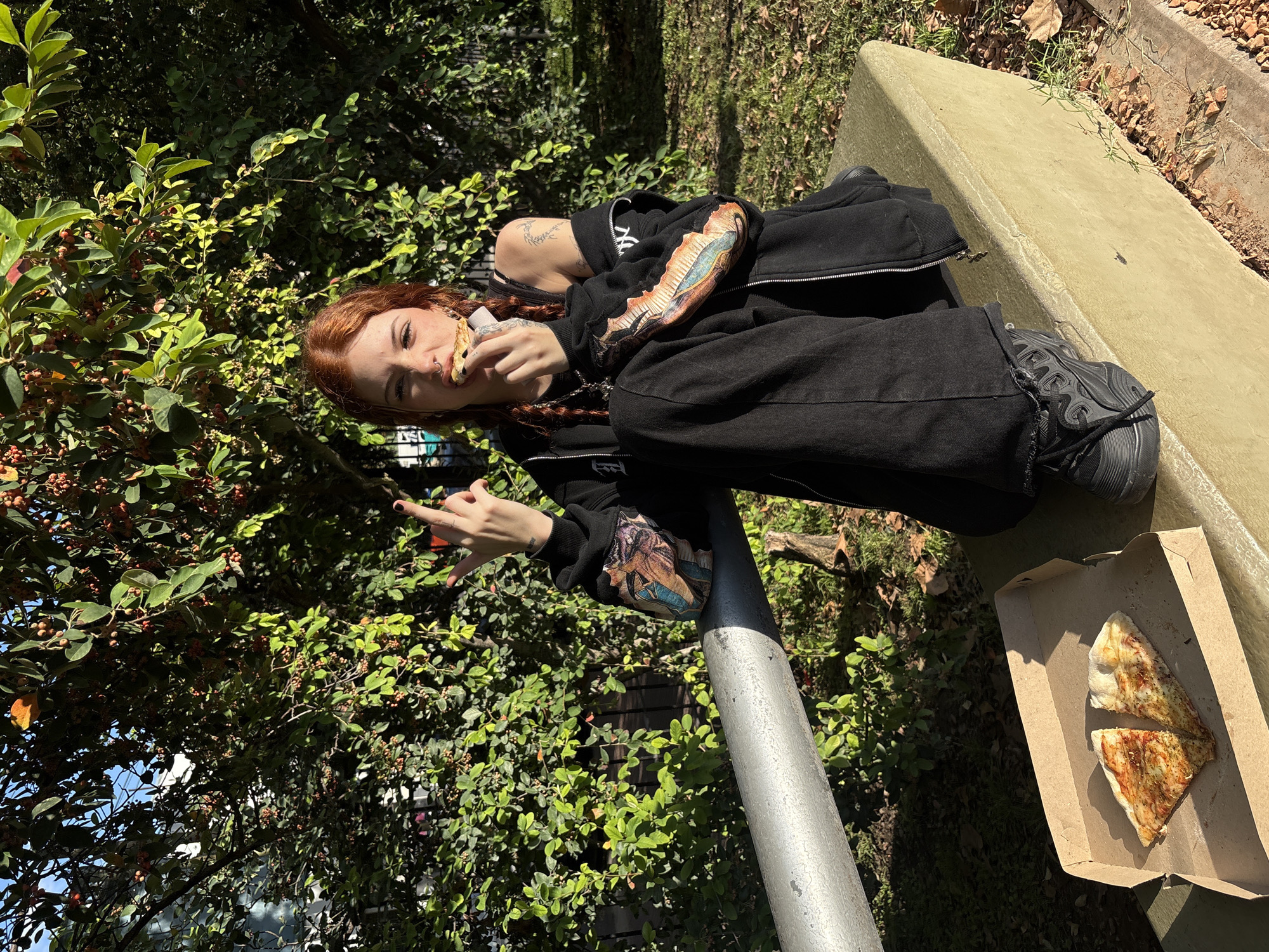 A young woman with red hair is eating pizza outside on a sunny day. She is sitting on a low, concrete wall next to a metal pole, surrounded by lush green trees and foliage. She is wearing a black hoodie with detailed artwork on the sleeves, black pants, and black sneakers. She is holding a slice of pizza in one hand and making a hand gesture with the other. An open pizza box with two remaining slices sits next to her on the wall. The setting appears to be a park or green space in Buenos Aires, Argentina, based on the local flora.