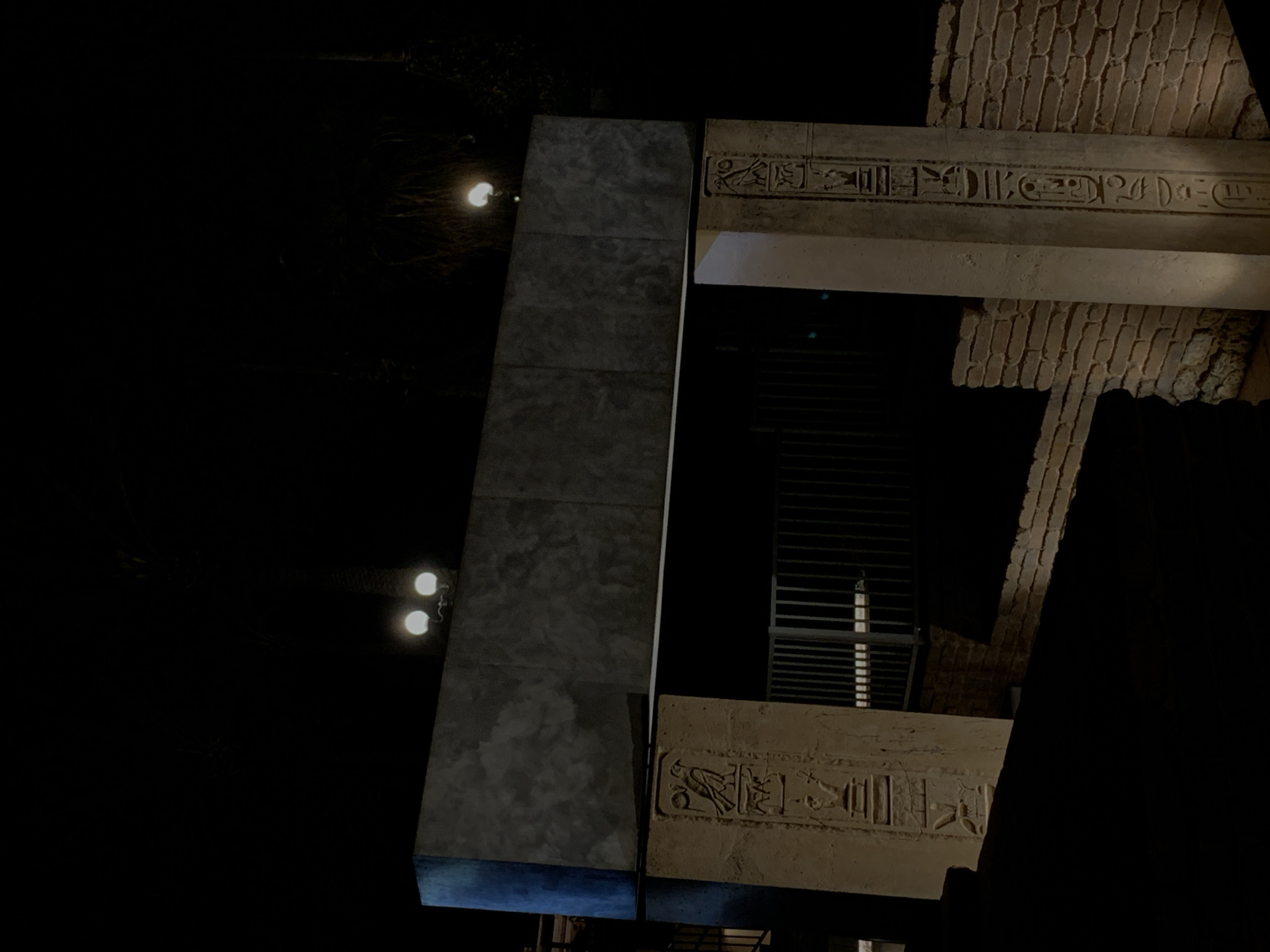 The image depicts an architectural structure in Tel Aviv, Israel, featuring ancient Egyptian motifs. The structure includes a large, rectangular column with a textured surface, possibly concrete or stone. Adjacent to the column are horizontal beams adorned with hieroglyphic inscriptions. A section of brickwork is visible, suggesting a larger construction. A building with horizontal blinds forms the background. Lighting fixtures cast a glow on the scene.