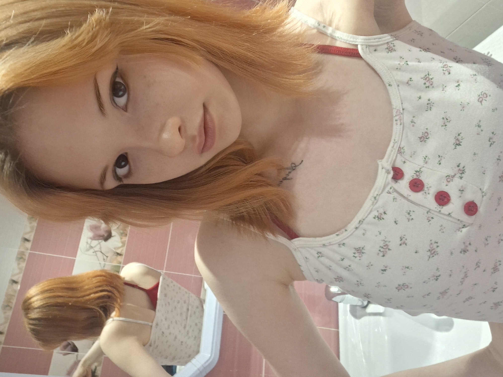 A young woman with reddish-blonde hair is posing for a selfie in what appears to be a bathroom. She is wearing a white tank top with a floral pattern and red buttons. The bathroom has pink tiles and a white sink. A mirror reflects the woman and the bathroom scene behind her. The location is assumed to be in the city of Chaikovsky, Russia.