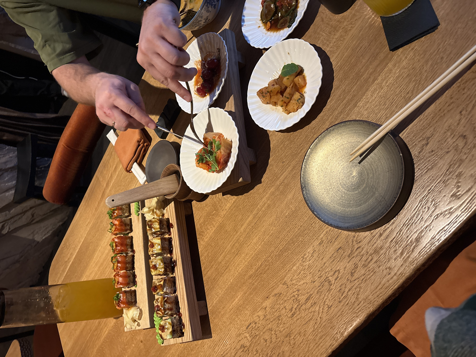 A top-down shot shows a partially visible individual seated at a wooden table. The individual is wearing a green shirt and a watch is visible on their wrist. They are using cutlery to select food from a white plate containing orange and green garnish. On the table are several other white plates containing various appetizers: one with dark-colored pickled vegetables, another with brown cubed food topped with a green leaf, and one with dark red cherries in sauce. There is also a long wooden serving dish filled with sushi rolls and a glass of orange liquid nearby. A small brown object, possibly a wallet, sits near the edge of the table. Chopsticks rest on a small, dark grey dish. The image evokes a dining scene, possibly in a restaurant in Krasnoyarsk, Russia.