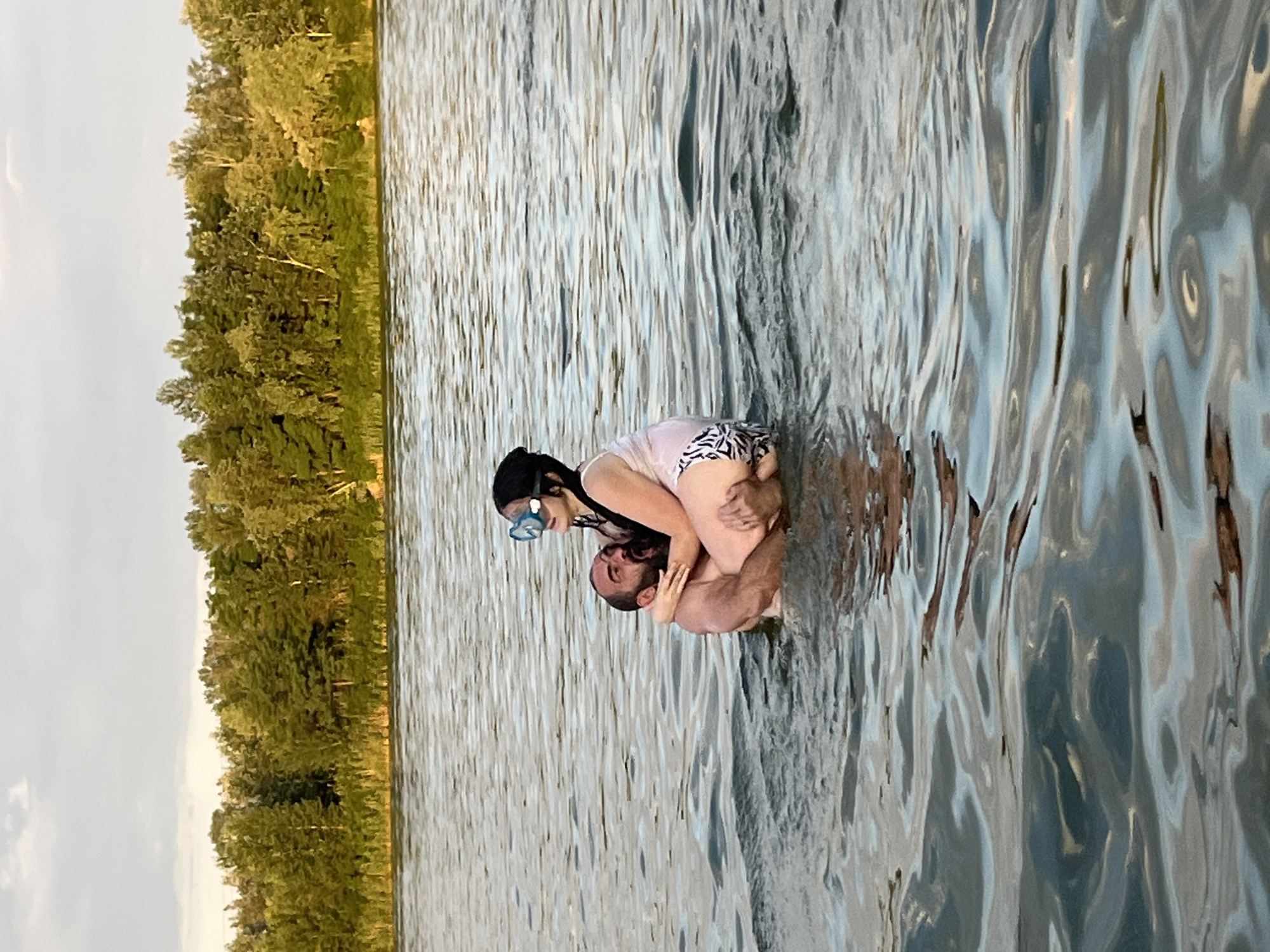 The image, oriented vertically, shows two people in the water with a treeline in the background. A man with a beard is being embraced by a woman who is wearing a black headscarf and goggles, suggesting she is either swimming or snorkeling. She is also wearing a white and black patterned swimsuit. The man is wearing swim trunks. They appear to be in a playful or affectionate pose.
The setting is a body of water, possibly a lake or river, with a dense line of trees visible along the bank. The water is calm with some ripples and reflections on its surface. The sky is overcast, indicating it might be early morning or late afternoon, or simply a cloudy day. The overall impression is one of a relaxed, natural setting, likely a vacation spot or a leisurely outing. The location context suggests this scene is in Dnipro, Ukraine. There is no visible text in the image.
