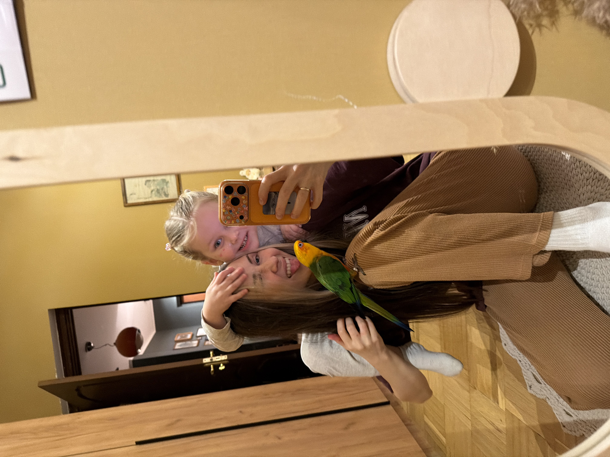 A mirror selfie shows a woman, a child, and a bird in a room. The woman is holding the phone, with a child leaning in close to her. The bird is perched on the woman's shoulder. The room contains a wooden wardrobe, a door, and a light fixture visible in the mirror. There is no evidence to support the location being in Khimki, Russia.