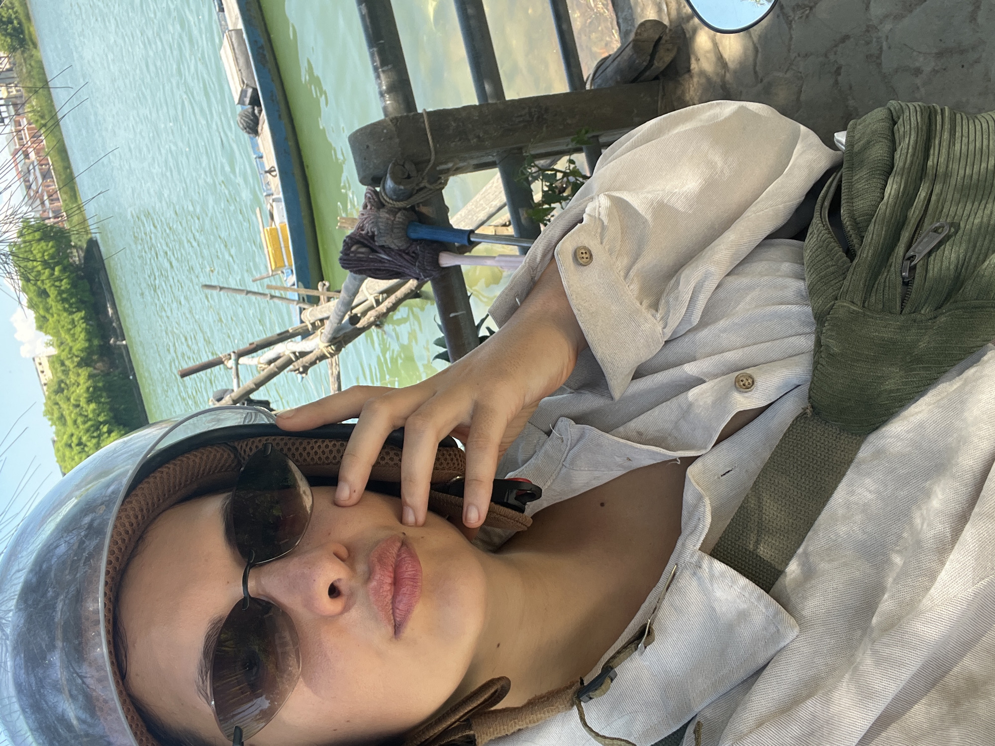 A person wearing a helmet and sunglasses pouts for a selfie in front of a body of water. The person is wearing a light-colored button-up shirt and a green fanny pack. In the background, there is a small blue boat with yellow crates, a wooden fence, and trees. The scene appears to be in a rural or waterfront area, possibly a canal or river, with evidence of a roadway or path in the foreground where the selfie is being taken. The water is calm and a light green color, reflecting the sunny sky.