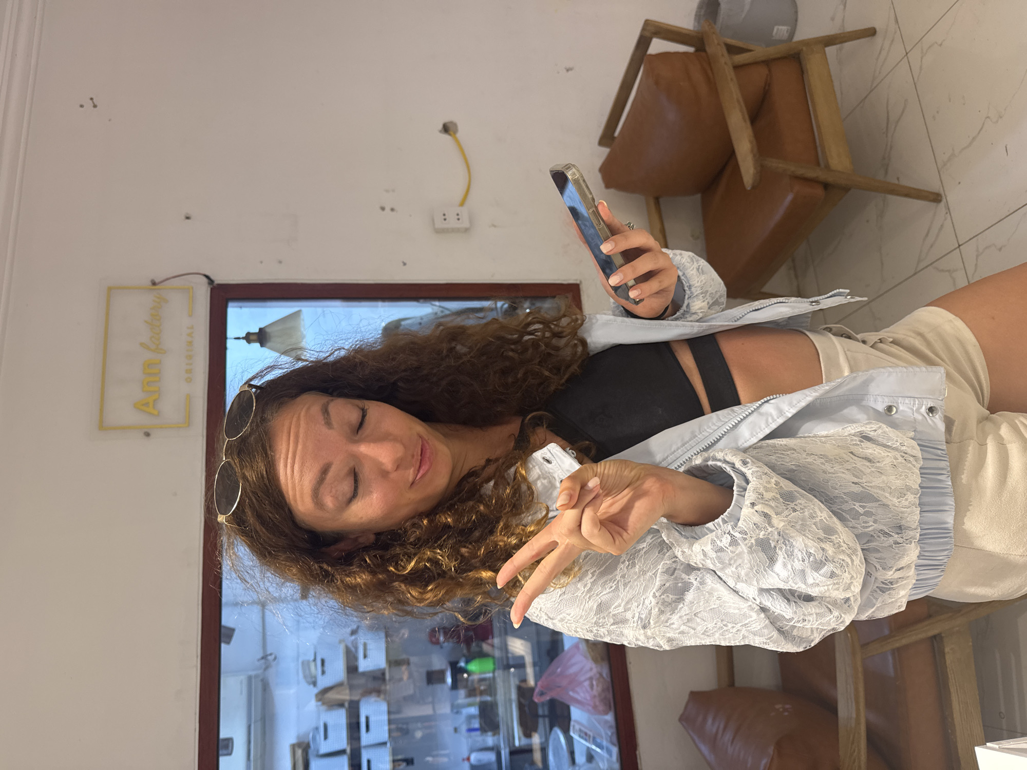 A woman with curly brown hair and sunglasses perched on her head is standing indoors and looking at her phone. She is wearing a light blue lace jacket over a black top and light-colored shorts. Her left hand is raised, displaying a peace sign, while her right hand holds a smartphone. In the background, there is a sign that reads "Ann factory ORIGINAL" and two brown leather chairs. The location is identified as Mộc Châu, Vietnam.