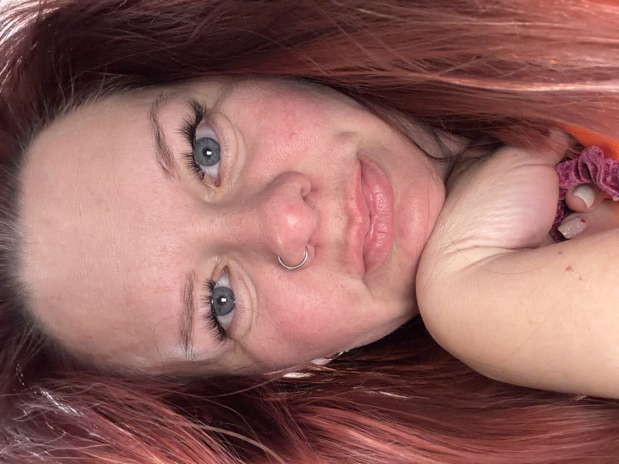 A close-up portrait of a fair-skinned woman with light blue eyes and pink hair. She has a silver nose ring. The image is oriented sideways, with the woman's head tilted to the left. She appears to be holding her right arm up near her face. A pink scrunchie is partially visible near her hand. The location cannot be determined from the image.
