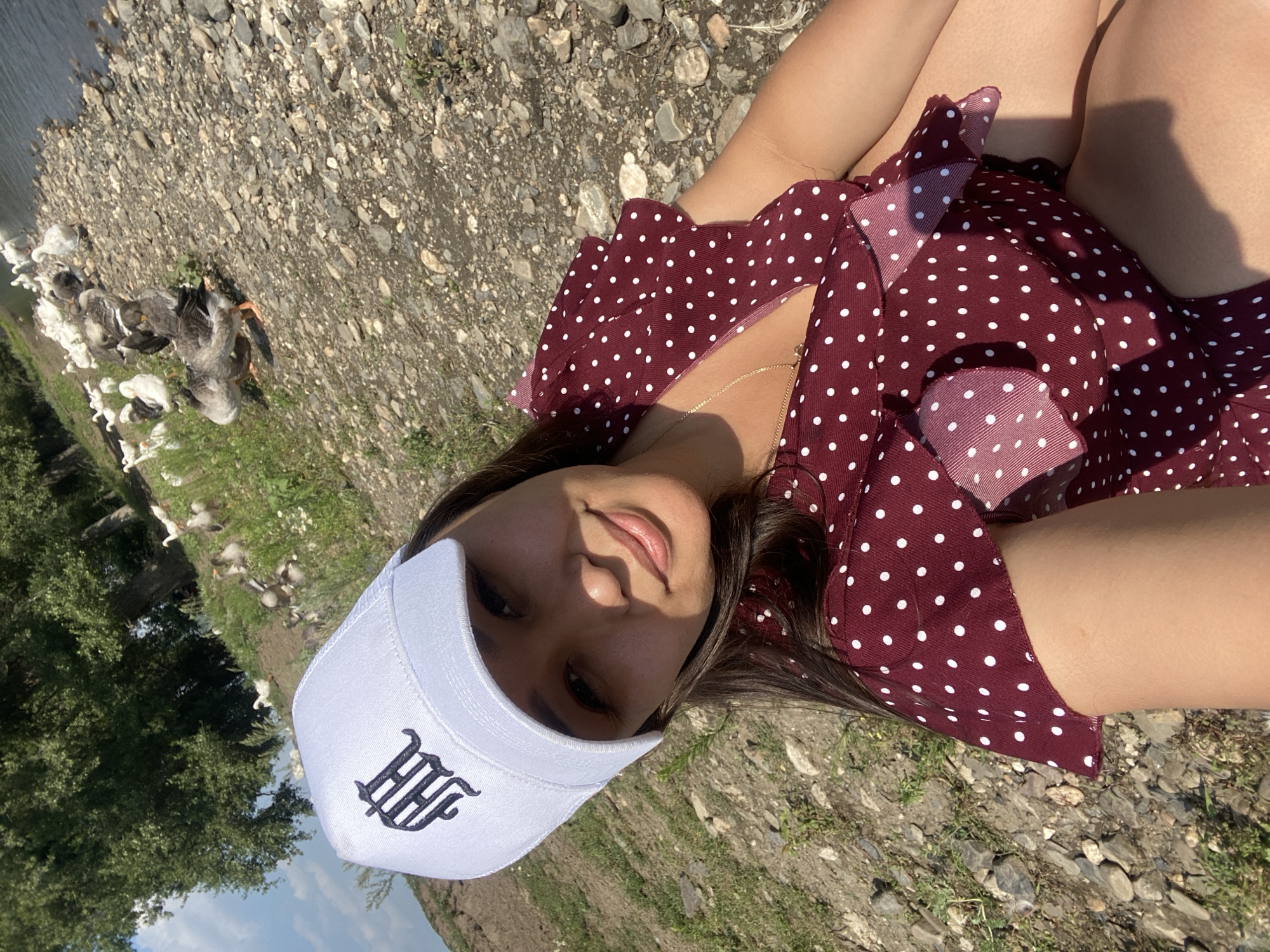 A young woman lies on the ground, her head tilted to the side. She wears a white baseball cap with a black, ornate logo and a maroon polka dot dress. Her eyes are open, and she smiles faintly at the camera. Sunlight casts a shadow on her face from her cap. In the background, a body of water is visible, and a flock of geese waddles along the rocky shore. Green trees and vegetation border the water. The scene appears to be set outdoors on a sunny day, possibly in a rural area like Maloarslangulovo, Russia. The overall mood seems relaxed and cheerful.