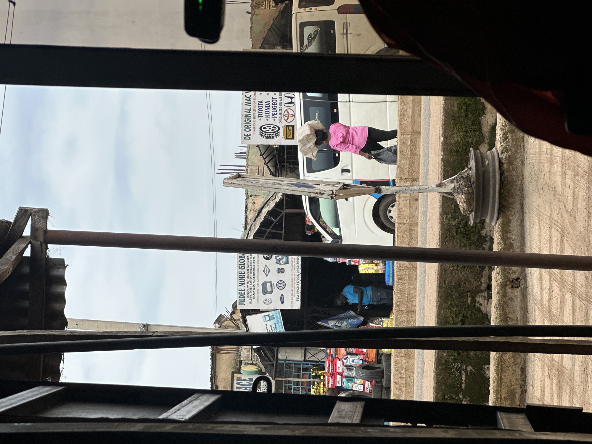 The image captures a street scene in Bukuru, Nigeria, viewed from a semi-enclosed space, likely a vehicle or shop entrance. In the foreground, dark vertical bars and a window frame obstruct the view, suggesting the perspective is from inside. The middle ground reveals a street with a raised concrete barrier. On this barrier, a person in a bright pink shirt and black pants carries a large white bundle on their head and a black bag in their hand. They are walking past parked white vans. Behind the vans, various shops are visible, including one with a sign for "JUDEE MORE GLOBAL," listing auto parts and mentioning brands like Toyota, Honda, and Peugeot. Another sign, "DE ORIGINAL MAC," also advertises auto parts. Stacks of tires and other automotive accessories are displayed outside some of the shops. The environment appears to be a commercial area with evidence of ongoing activity.