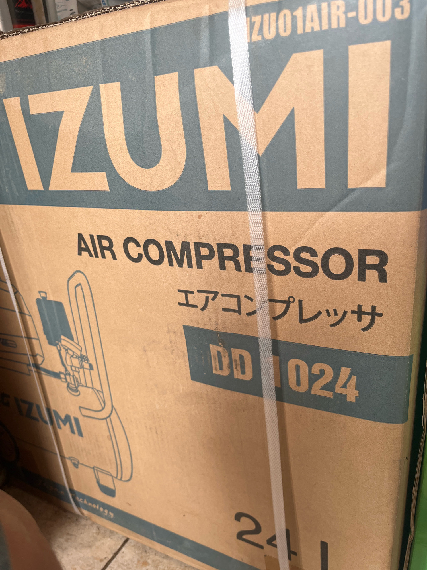 The image shows a cardboard box for an IZUMI air compressor. The box is light brown with dark blue text and graphics. The brand name "IZUMI" is prominently displayed in large, light brown letters. Above the brand name, "ZU01AIR-003" is printed in smaller, dark blue letters. Below "IZUMI", the words "AIR COMPRESSOR" are printed in dark blue. Further down, Japanese characters "エアコンプレッサ" are also printed in dark blue. A model number "DD 1024" is visible in light brown letters on a dark blue background. A graphic of an air compressor is drawn on the left side of the box. Near the bottom of the box, "IZUMI" is again printed in dark blue, and "24L" is visible in large numbers. The box is bound with a white plastic strap. The background suggests the box is indoors, possibly in a storage or retail environment, with a tiled floor visible at the bottom. The image's lighting is somewhat dim and angled, creating shadows. The location in Karang Sari, Indonesia is not depicted in the image.