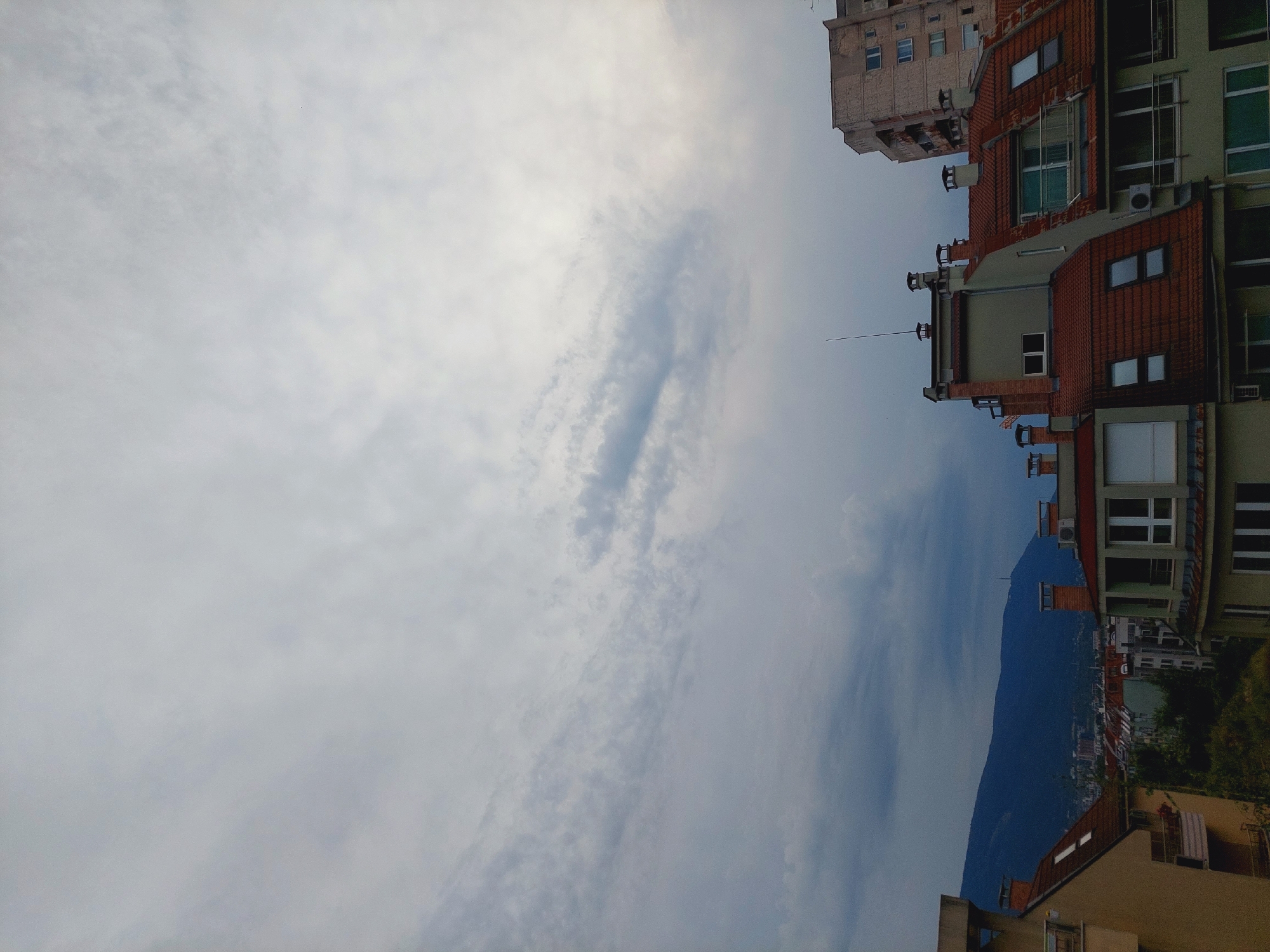 The image shows a view of rooftops and a mountain in Sofia, Bulgaria, under a cloudy sky. In the foreground, several buildings with terracotta tiled roofs are visible, featuring chimneys and windows. To the left, part of a yellow building with balconies can be seen. In the background, a mountain range, likely Vitosha, dominates the horizon. A few smaller buildings and trees are scattered between the foreground rooftops and the mountain. The sky is overcast with varying shades of gray clouds, suggesting a potential for rain or an approaching storm. No people or animals are discernible in the image.
