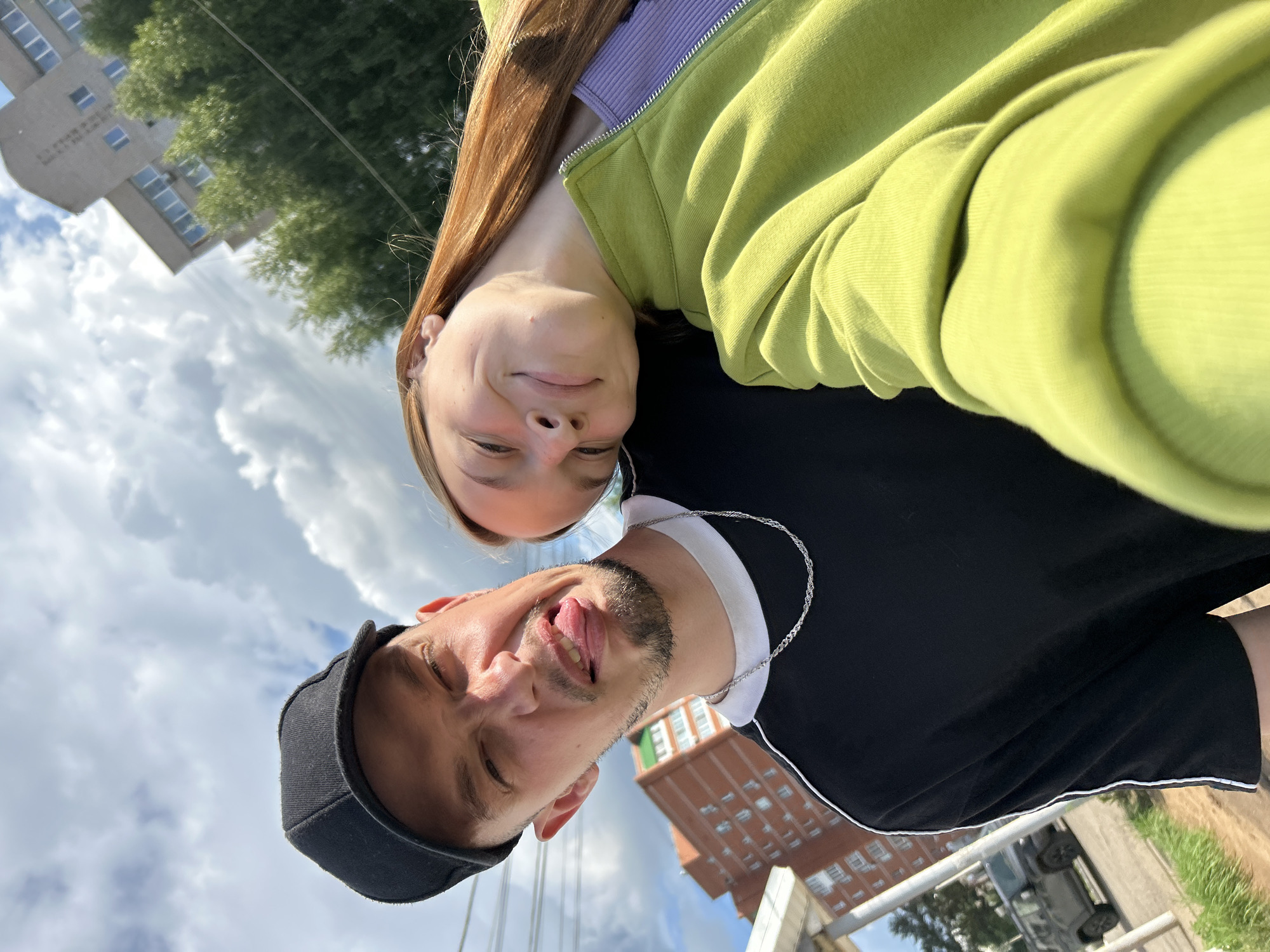 A selfie taken outdoors on a cloudy day in Chaikovsky, Russia, features a man and a woman. The man, in the foreground, wears a black baseball cap and a black t-shirt with white piping. He is sticking his tongue out and making a face. A silver chain is visible around his neck. The woman is leaning her head on the man's shoulder, wearing a light purple top and a lime green zip-up jacket. Her eyes are closed, and she has a slight smile. In the background, apartment buildings, trees, and power lines are visible. A dark-colored SUV is parked on the left side of the frame. The overall setting suggests a residential area of the city.