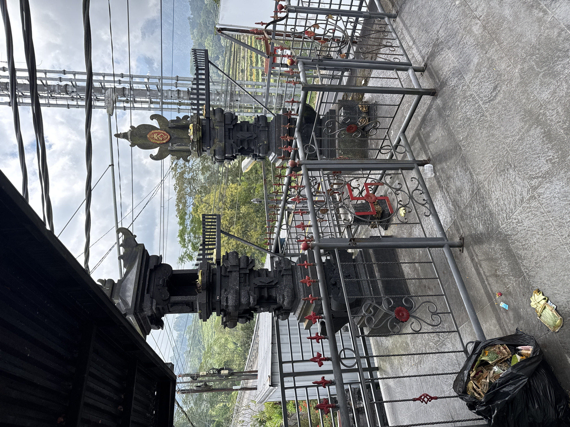 The image shows a view in Baturiti, Indonesia. Two black, ornate stone structures stand behind a gray metal fence with red accents. Overhead, electrical wires crisscross the sky. In the background, a hillside with terraced fields and vegetation is visible. A black trash bag full of debris sits on the gray tiled ground near the fence. Scattered pieces of trash lie nearby.