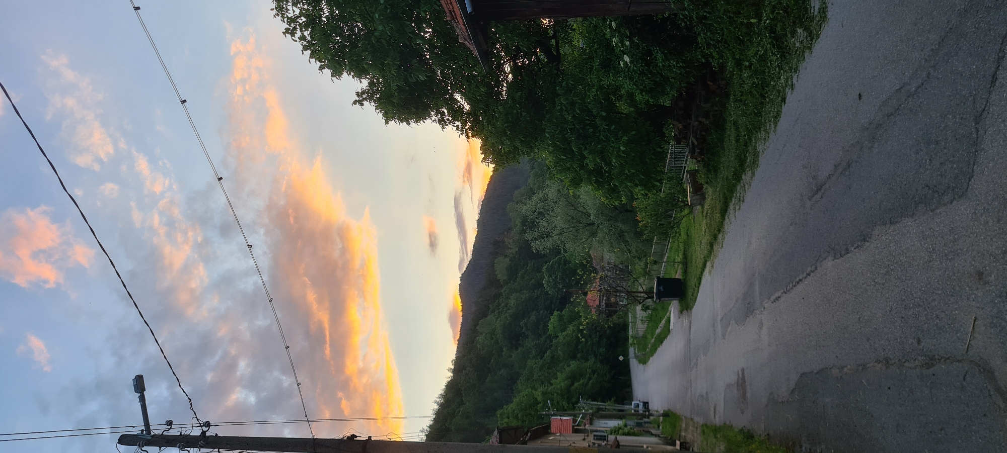 This image depicts a quiet, rural village street in Lopian, Bulgaria, at dusk or dawn, characterized by a vibrant sunset/sunrise sky.
The upper half of the image features a sky with scattered clouds, displaying a gradient of colors from pale blue at the top to intense orange, yellow, and pink hues concentrated towards the horizon line above the mountains. Several dark, thin utility wires extend horizontally across the sky from a wooden utility pole positioned on the far left. A grey streetlamp arm protrudes from this pole. Small, dark specks, likely birds, are visible against the lighter parts of the sky in the upper right quadrant.
The midground is dominated by a dense, forested mountain range, its contours highlighted by the low-angle sunlight. The peak of the sun's glow is visible as an intense orange band just above the mountain ridge on the right side of the frame. The trees covering the mountains are a mix of dark green evergreens and lighter green deciduous varieties.
The foreground and lower half of the image show an asphalt road that curves gently uphill and to the left, receding into the distance. The road surface appears worn, with visible patches and darker, damp areas. On the left side of the road, several older, multi-story houses with orange-tiled roofs and light-colored, somewhat weathered facades are visible. A small, light-colored vehicle is parked on the side of the road near these houses. The wooden utility pole from the top left extends down into this section.
On the right side of the road, a grassy verge runs parallel to a white metal picket fence. Various trees and shrubs, including a large, full deciduous tree with bright green leaves, are situated along this side. A dark blue outdoor trash bin is visible next to the fence. Further down the road on the right, a neatly stacked pile of firewood is present, along with the corner of a building featuring a red-tiled roof and dark wooden eaves. No individuals are visible, and no discernible actions are occurring within the frame.