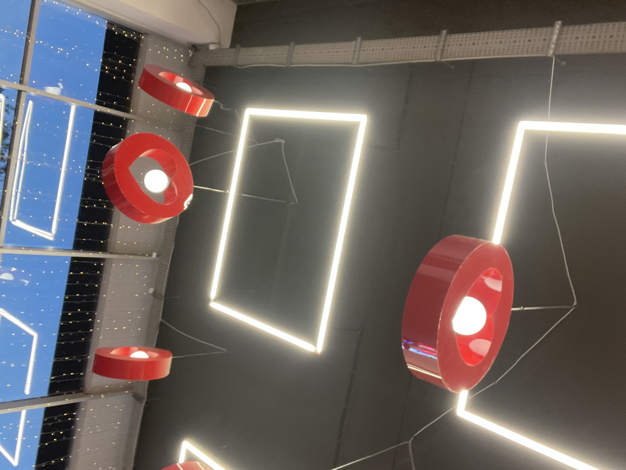 The image shows a ceiling with modern lighting fixtures and decorative elements. There are several rectangular and square-shaped LED light panels emitting a bright white light, suspended from the dark ceiling. Hanging below these panels are red, heart-shaped pendant lights, each with a visible white light bulb inside. In the background, there are large windows that appear to look out onto a bright blue sky, possibly indicating daytime. Along the top of the windows, there are strings of small, yellow fairy lights. To the left of the image, a metallic conduit runs vertically along the ceiling. The overall impression is of a modern, possibly commercial or entertainment space, decorated with a festive or romantic theme.