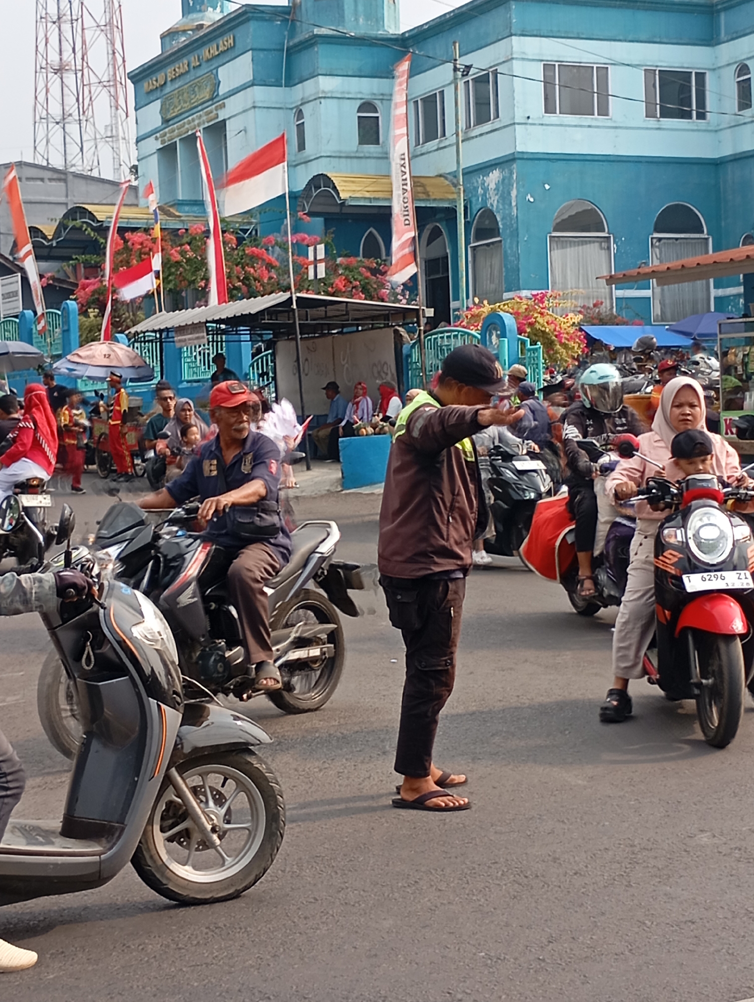 The image depicts a street scene in Subang, Indonesia, with a prominent blue mosque in the background, identified by the inscription "MASJID BESAR AL-IKHLAS". The street is busy with motorcycle traffic. In the foreground, a man in a brown jacket and dark pants is standing on the road, extending his right hand as if directing traffic. He is wearing dark flip-flops. To his right, a woman wearing a light pink headscarf and a light pink jacket is riding a red scooter with a child wearing a black cap in front of her. The scooter's license plate is visible, reading "T 6296 ZL". Further in the background, more motorcycles and people are visible. Several Indonesian flags are displayed on poles, fluttering in the wind. The environment appears to be an urban setting with a mix of vehicles and pedestrians.