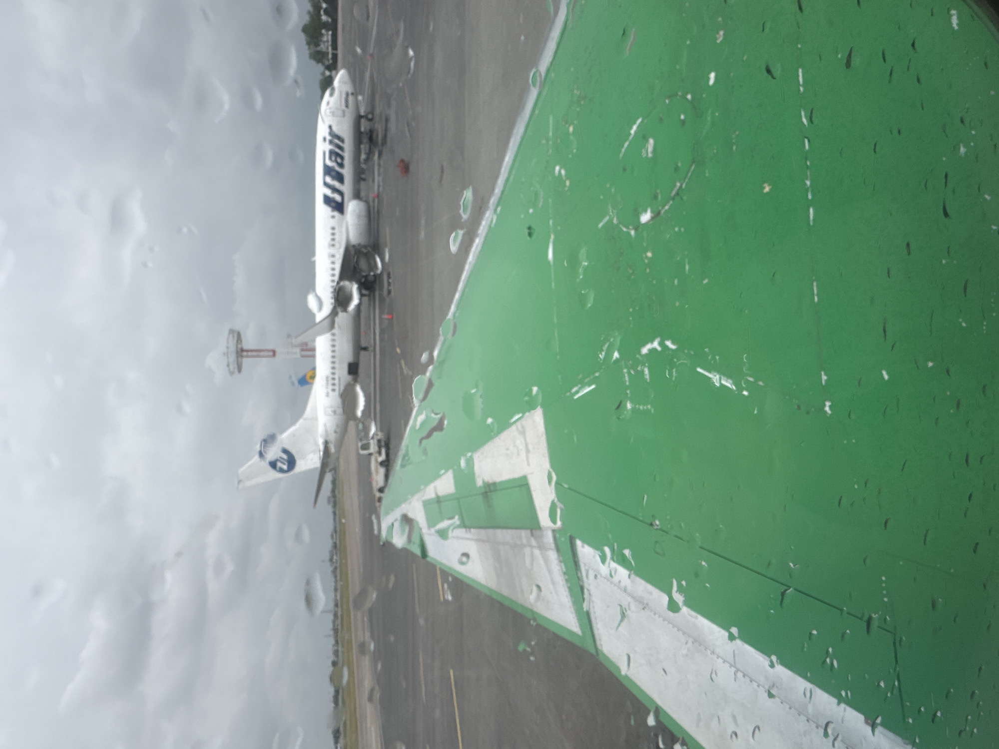 The image shows a UTair airplane parked on a wet tarmac. Raindrops are visible on the window through which the photo was taken. The wing of the aircraft, painted green with white markings, is prominently displayed in the foreground. The image was taken in Tashkent, Uzbekistan.
