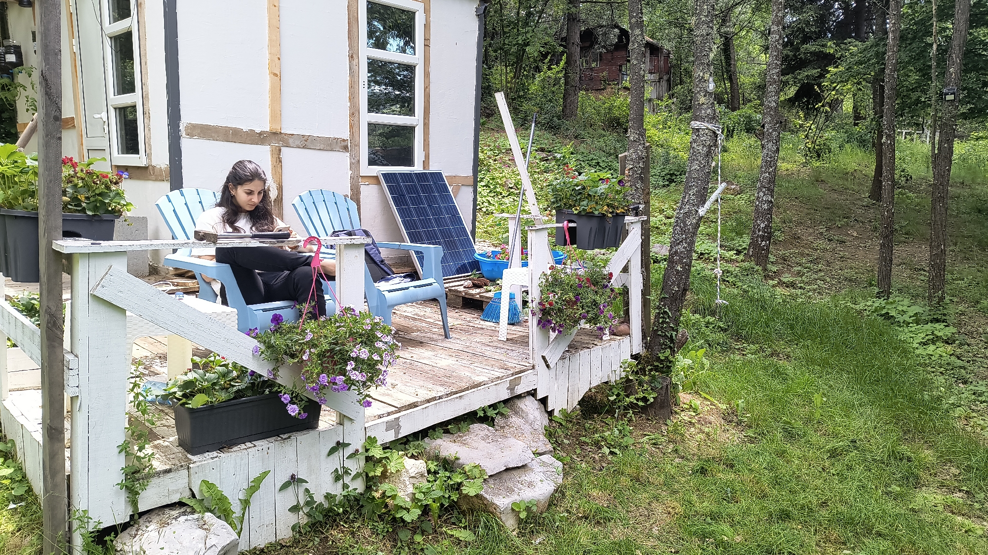A young woman with long dark hair is sitting on a light blue Adirondack chair on a wooden porch. She is wearing a white long-sleeved shirt and black pants, and she is looking down at a device in her hands. The porch is white and weathered, with purple flowers in hanging baskets and planters. To the left of the woman, there is a solar panel leaning against a white structure with large windows. Behind the porch, there is a lush green forest with trees and other vegetation. In the distance, a small wooden building is visible. The scene appears to be a rural or remote location, possibly a cabin or a small house in the woods. The image does not provide specific details that would indicate the location in Dramsha, Bulgaria.