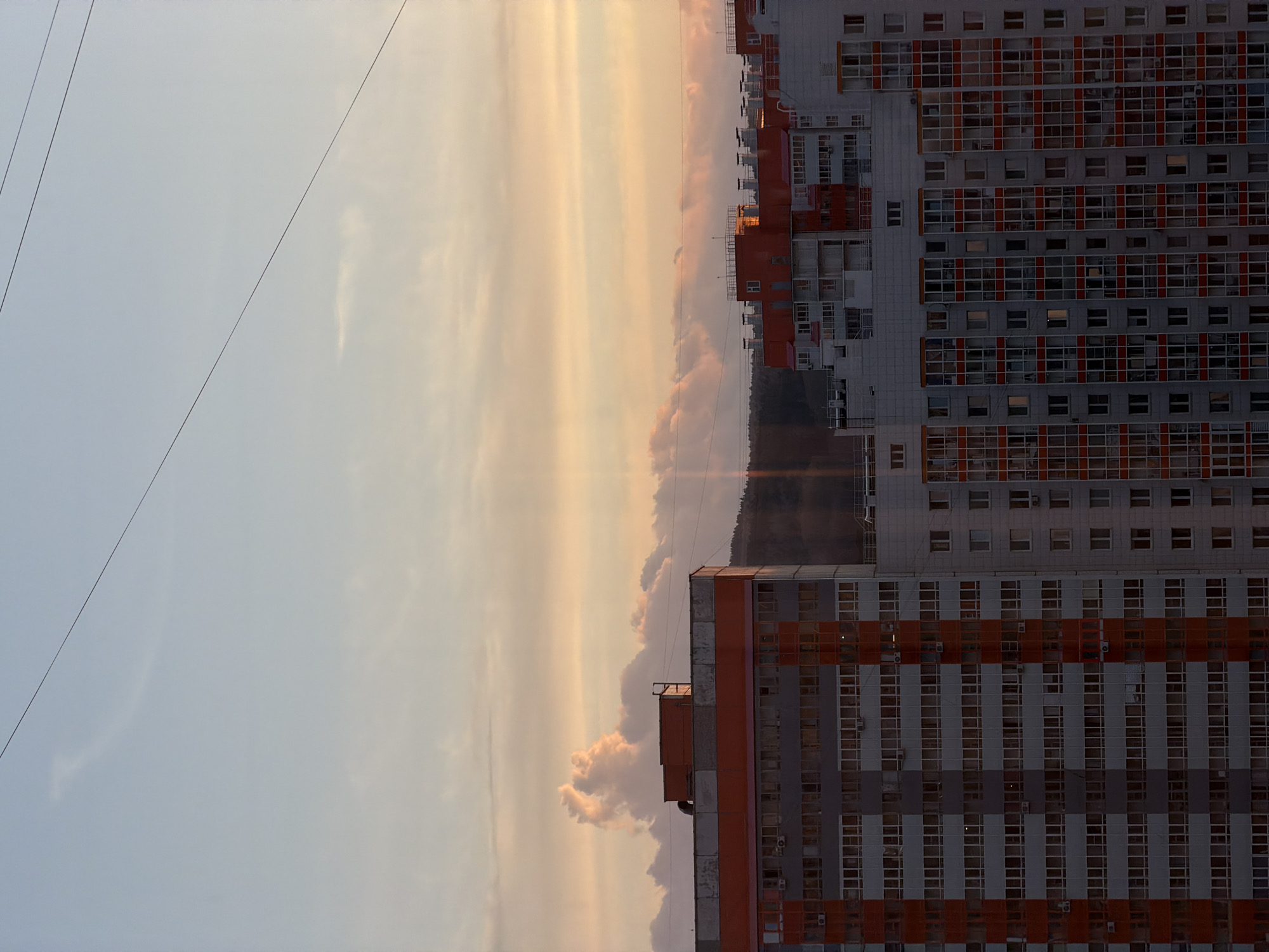 The image shows a tall, grey apartment building with red accents, located in Krasnoyarsk, Russia. The building is densely packed with many rectangular windows. Power lines are visible in the upper left corner. In the background is a sky with streaks of orange, pink, and grey clouds, suggesting a sunset or sunrise. There also appears to be a treeline separating the building from the sky.