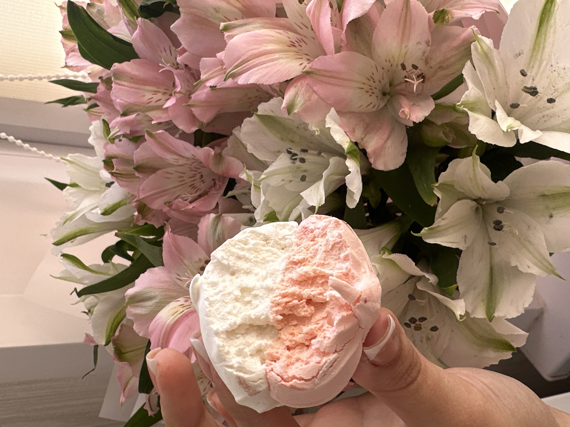 An adult's hand, exhibiting manicured nails with white tips, holds a bicolored dessert item. The dessert, likely a marshmallow or zephyr, is approximately spherical and shows a clean break or bite revealing an internal texture consistent with aerated confectionery. The upper portion of the dessert is white, while the lower portion is pinkish-orange. The hand and dessert are positioned in the foreground, obscuring a portion of a densely arranged bouquet of flowers. The bouquet consists primarily of pink and white alstroemeria (Peruvian lilies) with prominent green foliage and visible anthers. The background is a light-toned, indistinct surface, possibly a wall or window, with a thin, light-colored beaded cord visible vertically on the right side, suggesting an indoor setting. The specific location within the city of Chaikovsky, Russia, cannot be determined from the visual evidence presented.