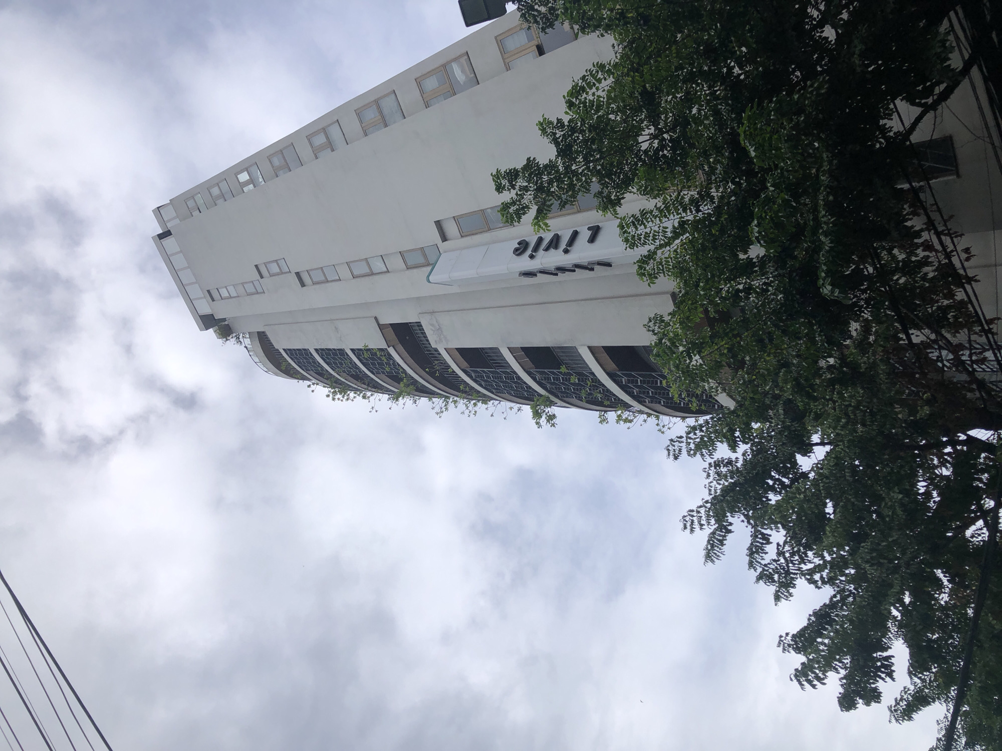 The image shows a tall, light-colored building in Đà Nẵng, Vietnam, with a grey, cloudy sky. The building has a modern, curved design with multiple floors and balconies filled with greenery. The name "Livie" is prominently displayed in black lettering on a white vertical sign on the side of the building. There are trees in the foreground, obscuring the lower part of the building. Overhead, thin wires crisscross the sky. The perspective is from a low angle, looking upwards at the building.