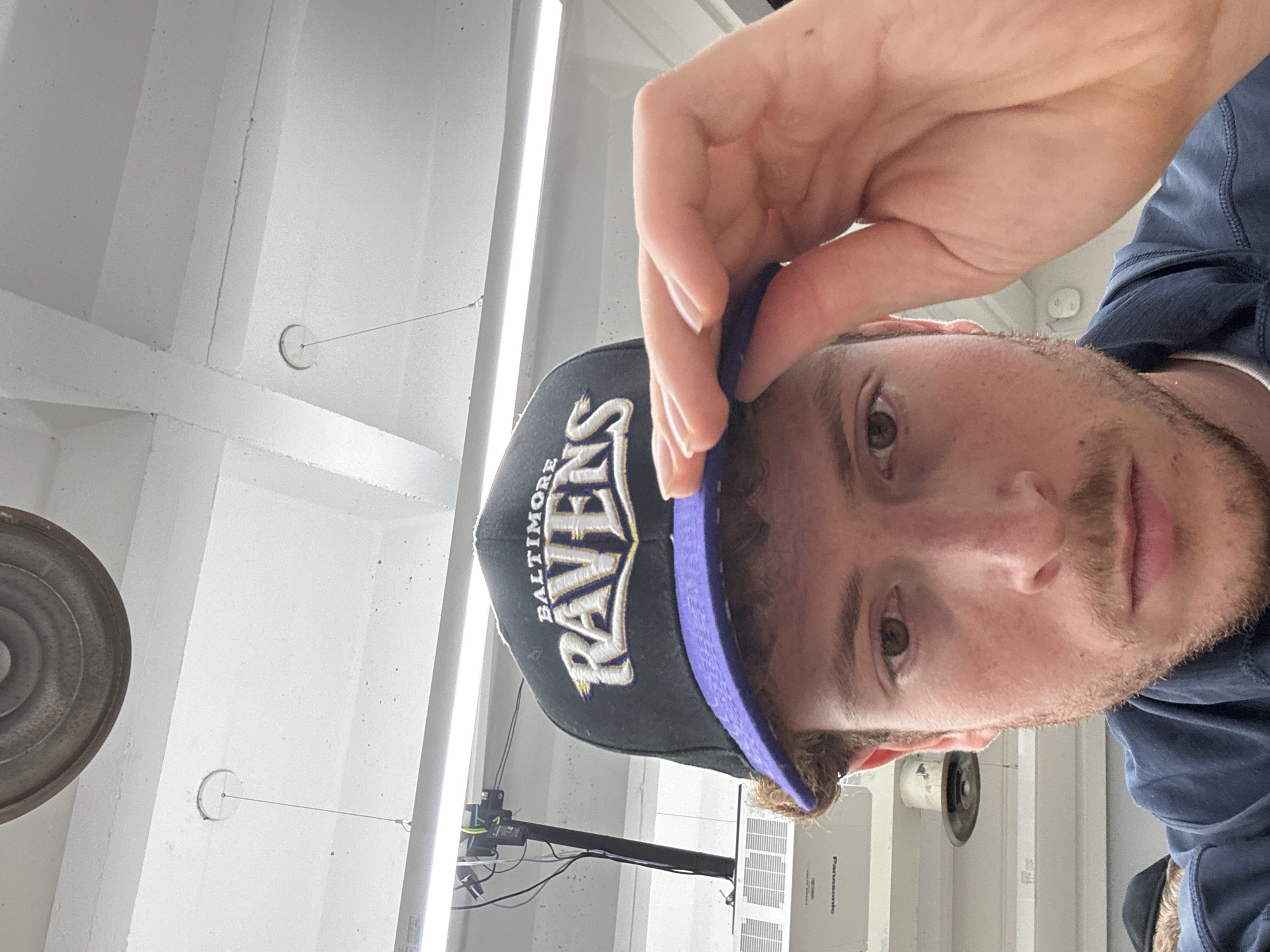 A young man is captured in a selfie, looking directly at the camera with a neutral expression. The image is rotated, presenting the man's head and shoulders from a low angle. He has light brown hair, stubble around his jawline and chin, and brown eyes. He wears a dark blue long-sleeved shirt and a black baseball cap. His right hand is seen holding the purple brim of the cap.
The cap prominently displays "BALTIMORE RAVENS" in white and purple embroidered letters on the front.
The background is an indoor, brightly lit space with white walls and a white ceiling featuring exposed rectangular beams. Long, linear fluorescent-style light fixtures run across the ceiling. Various objects are visible in the background: a large, circular metal disc or vent on the left, a smaller circular object with a wire extending from it, and what appears to be a wall-mounted air conditioning unit or projector. The word "Panasonic" is partially discernible on this unit. Wires and cables are also visible against the white walls. The overall impression is a clean, possibly industrial or modern office/studio environment.