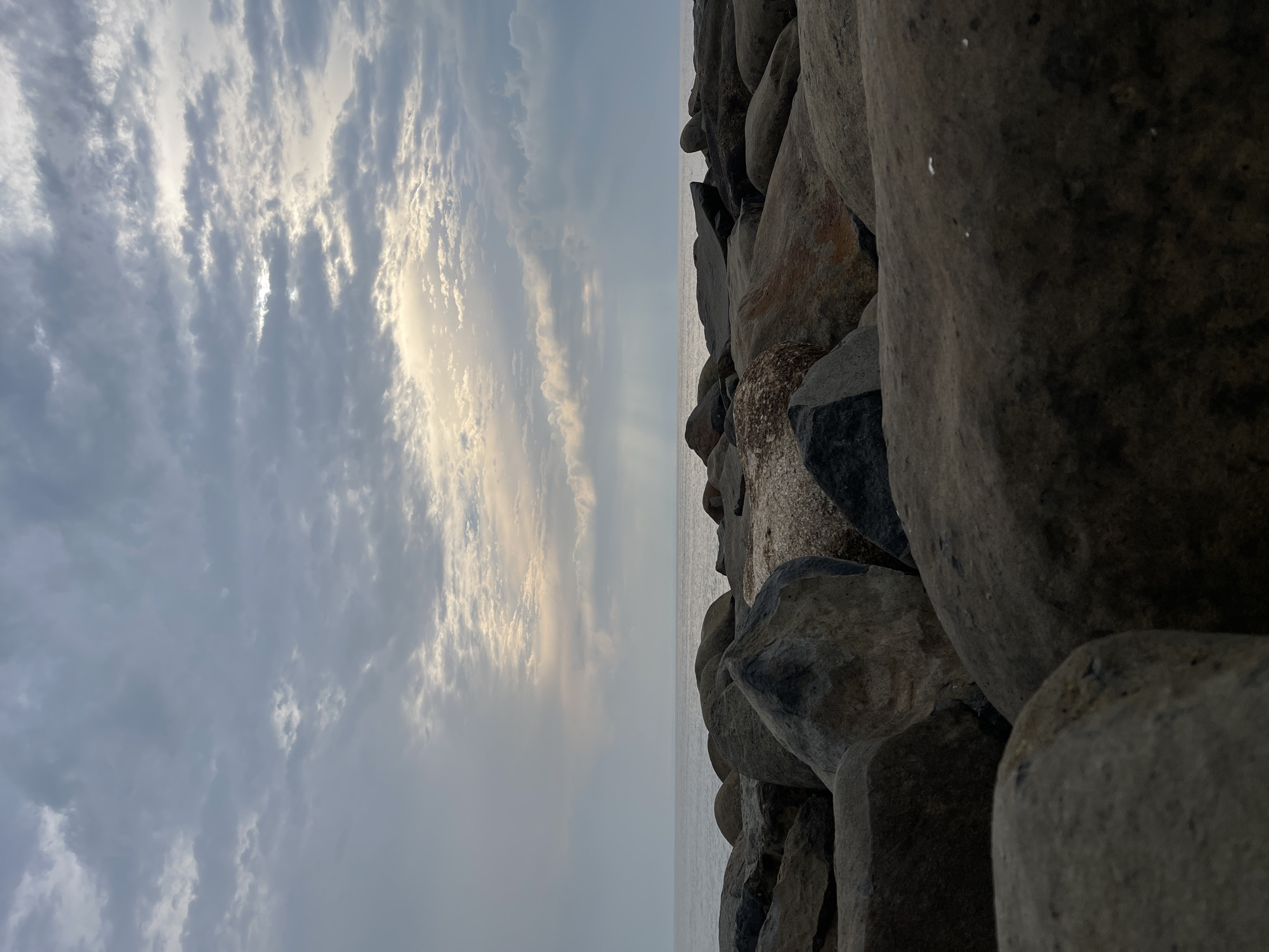 A low-angle, wide shot captures a rocky shore leading to a calm body of water under a cloudy sky. The foreground is dominated by large, grey, and tan rocks of various shapes and sizes, some appearing wet. The middle ground features the tranquil sea, meeting the horizon under a diffused, muted light. The sky is filled with a textured layer of clouds, with patches of brighter sky and subtle sunbeams breaking through in the upper right quadrant. The overall impression is one of a serene, natural landscape, likely on the coast of Kualacangkoi, Indonesia, given the mention. There are no people or animals visible in the frame. The lighting suggests it is either early morning or late afternoon.