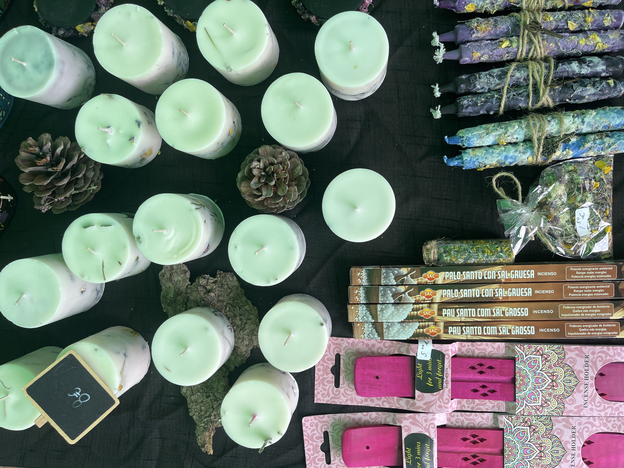 An overhead view captures a display of various products, predominantly candles and incense, laid out on a dark, likely black, fabric surface. The scene suggests a market stall in the city of Unknown, Unknown.
In the upper left quadrant, approximately seven light-green-topped pillar candles are arranged. Their lower bodies are lighter, appearing white or off-white, with visible embedded dried botanical materials such as petals and leaves. A small black chalkboard sign, handwritten with "8€" in white, leans against one of these candles.
Scattered across the central and upper right areas are approximately twelve additional light-green-topped pillar candles, identical in style to those on the left. Two large, brown pinecones are interspersed among these candles, serving as decorative elements.
In the lower left quadrant, multiple packages of pink wooden incense holders are visible. Each package features a decorative cutout design and printed text, including "Light for 3 min and forget..." and "INCENSE HOLDER." Some packaging displays a pink and teal mandala pattern. A handwritten white label displaying "5€" is attached to one package.
To the right of the incense holders, six vertically stacked boxes of "PALO SANTO COM SAL GROSSO INSCENSO" (Palo Santo with Coarse Salt Incense) by the brand SAC are displayed. The packaging features an image of white crystalline material. Portuguese text on the boxes describes properties such as "Potente energizador de Ambiente" (Powerful environment energizer).
In the lower right quadrant, three bundles of handmade, textured candles are present, tied with natural twine. Two bundles consist of two variegated blue and green candles, while the other four are variegated purple and dark gray/black. Small metal keys are visibly tucked into the twine bindings of these bundles. Adjacent to these candles, a small clear plastic bag filled with dried plant material (leaves and petals) is visible, bearing a handwritten price tag of "1.50€."
No individuals are present in the image.