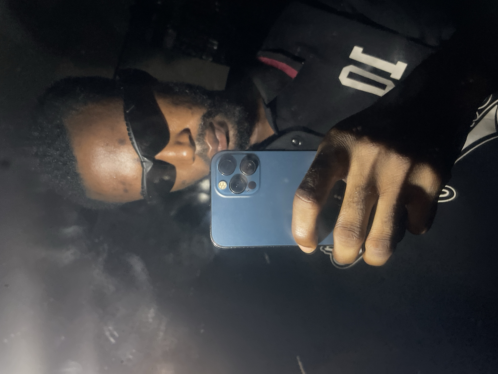 An adult male, with dark skin and a beard, is depicted in a low-light indoor setting within the city of Lokoja, Nigeria. The individual wears black wrap-around sunglasses and a black polo shirt featuring a red stripe detail on the collar/placket and the white numeric identifier "01" on the right chest area. He holds a blue smartphone, consistent with an iPhone model featuring a triple-lens camera system, in his right hand. The phone's screen is angled towards the subject, suggesting a self-portrait or screen interaction. A charging cable is connected to the phone's charging port. The background is dark and appears to be a reflective surface, possibly a window or mirror, showing blurred light reflections and specks.
