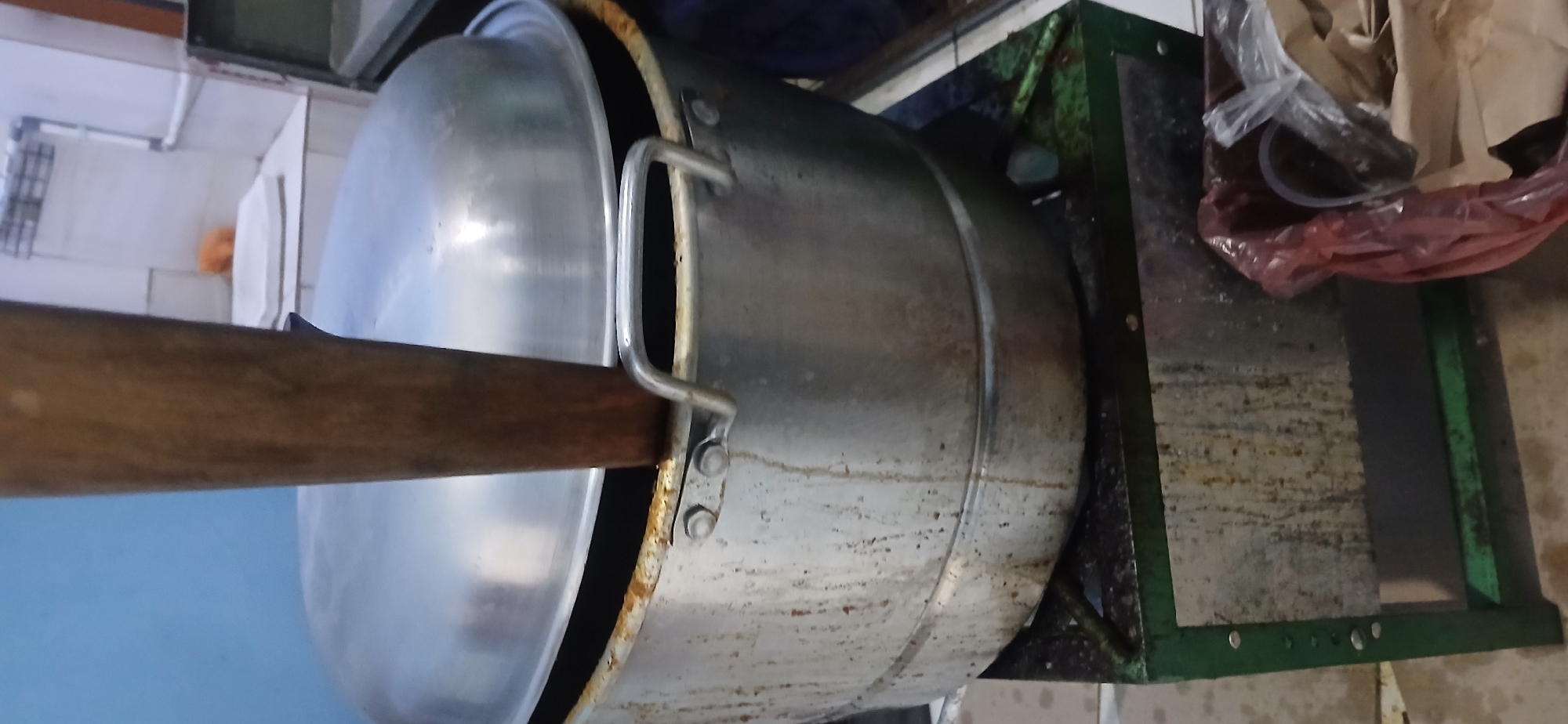 An over-the-shoulder, medium shot captures a large, aluminum pot with a wooden stirrer inserted into its lid. The pot rests on a green metal stove. The pot and stove are visibly aged and stained with grease and dirt, suggesting heavy use. In the background, a portion of a tiled wall and a sink are visible, with a hint of an orange object on the counter. To the right of the stove, a red plastic bag and a brown paper bag are piled on the floor. The image appears to be taken in a kitchen or food preparation area, possibly in West Jakarta, Indonesia, given the context provided. The scene conveys a sense of a functional, utilitarian space where food is prepared.