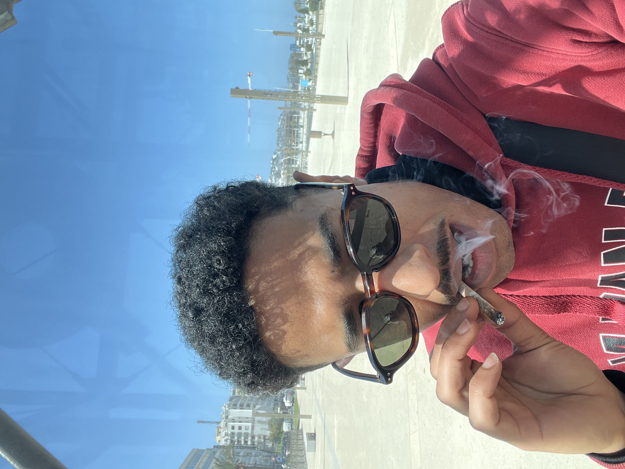 The image depicts a male individual in a close-up outdoor selfie, actively smoking a hand-rolled cigarette. The subject has dark, curly hair and a mustache, wearing tortoiseshell-patterned sunglasses with dark lenses and a red hooded sweatshirt over a black garment. His right hand holds the lit, hand-rolled cigarette near his mouth, from which visible plumes of white smoke are exhaling. The background reveals a sunny coastal urban environment in Tanger, Morocco. Visible elements include a wide, light-colored paved promenade, a sandy beach, and the sea on the right. The left side of the background shows modern, multi-story buildings, a street with parked vehicles, a round blue traffic sign, and a palm tree, all under a clear blue sky.
