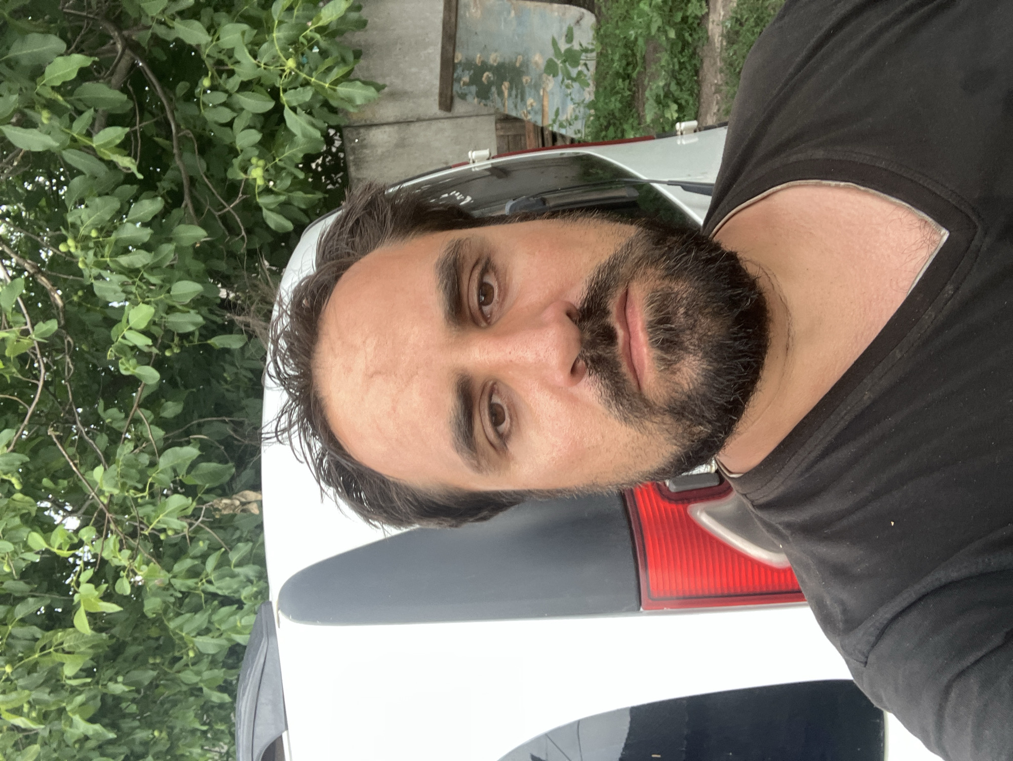 A man with dark hair and a beard is taking a selfie outdoors. He is wearing a black t-shirt with a v-neck. Behind him, to his left, is the white side of a vehicle with a dark grey accent. To his right, there is a concrete wall, some greenery, and stacked wooden planks. The background appears to be a rural or suburban environment, possibly in Vovkivka, Ukraine, given the presence of trees and what looks like a garden area. The lighting suggests it is daytime.
