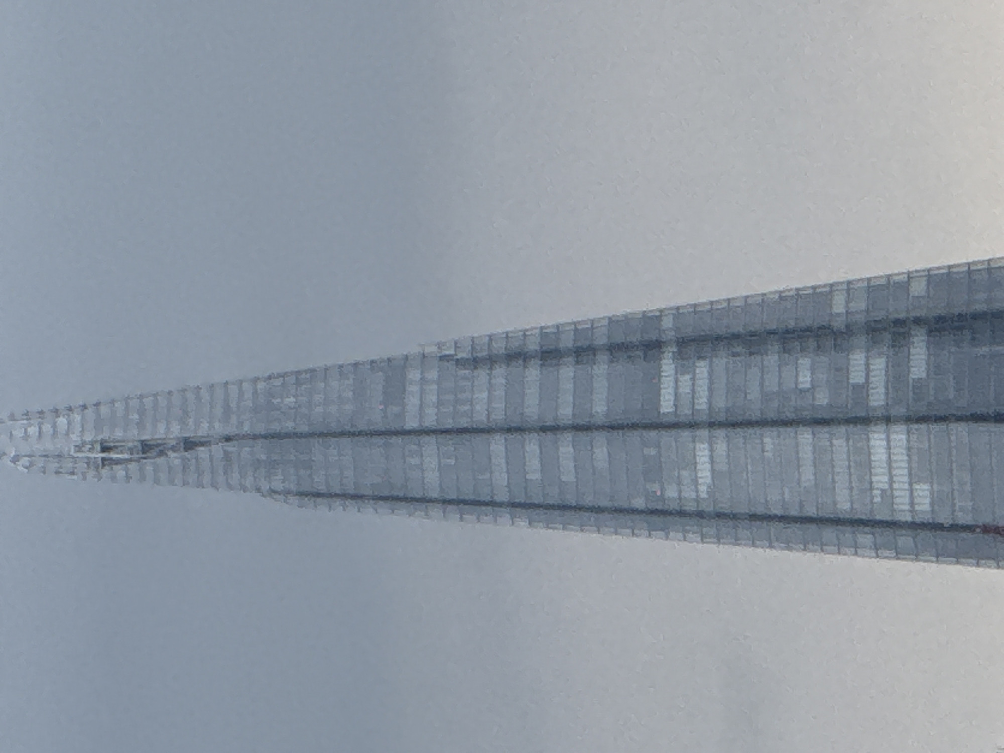 The image shows the top portion of The Shard skyscraper in London, United Kingdom. The building's glass facade is visible, receding into a hazy, overcast sky. The structure tapers to a point, its design characterized by sharp angles and reflective surfaces. The surrounding environment appears to be obscured by fog or cloud cover, muting the colors and reducing visibility.