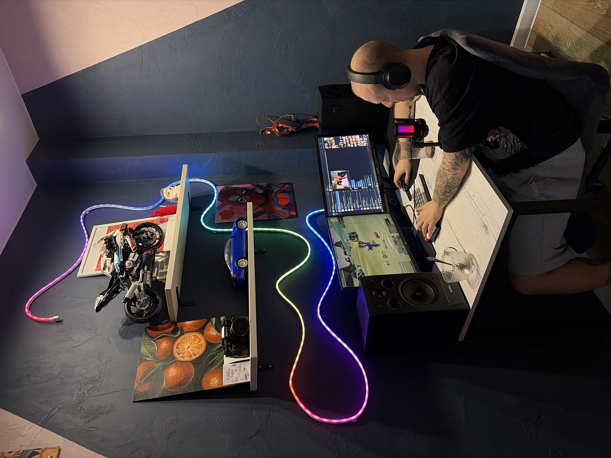 A man with a shaved head and headphones is seated at a desk with dual monitors in a room with dark blue walls. He is wearing a black t-shirt, grey shorts, and has tattoos on his arms. The top monitor displays what appears to be video editing software, while the bottom monitor shows a video game. To his left are two shelves mounted on the wall. On these shelves, there is a picture frame, a LEGO motorcycle model, a painting of oranges, a toy car, a Zenit camera, and a Spider-Man themed item. A multicolored LED strip light runs along the wall, connecting the shelves and monitors. A glass with a spoon inside is on the desk. The scene is captured from a high angle. The city is not visible in this image, and there is no information to suggest that the image was taken in Rostov-on-Don, Russia.
