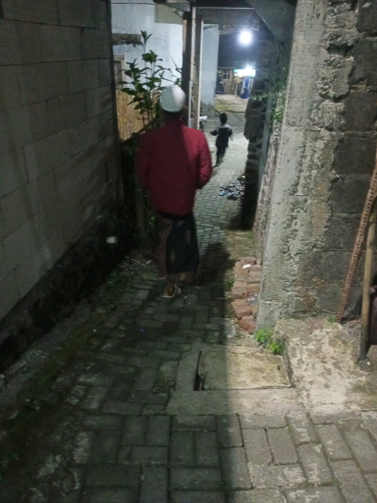 In Andongsili, Indonesia, at night, a person in a red jacket and a white kufi walks away from the camera on a paved pathway. The pathway is lined by rough, grey stone walls on both sides, with some overgrown greenery. A child in dark clothing is visible further down the pathway, running away from the camera. The scene appears to be a narrow alleyway between buildings, with artificial lights illuminating the area.