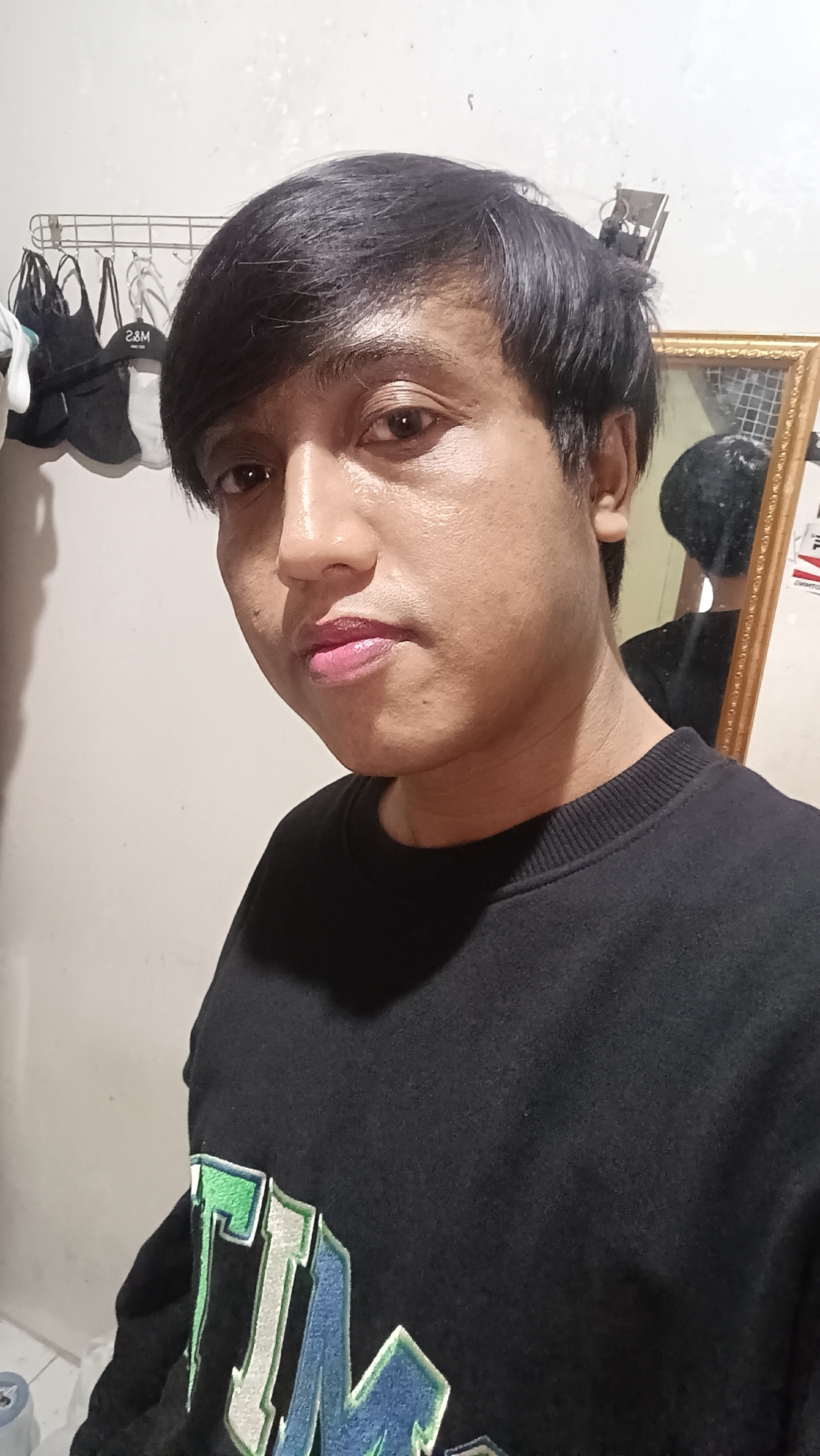 In Wangon, Indonesia, this selfie depicts a young person with dark hair and brown eyes, facing the camera directly. They are wearing a black sweatshirt with a graphic design on the front featuring the letters "TIM" in green and blue. The background shows a plain, light-colored wall, a metal rack holding several bags, and a gold-framed mirror reflecting the person's face and upper body. The lighting suggests an indoor setting.