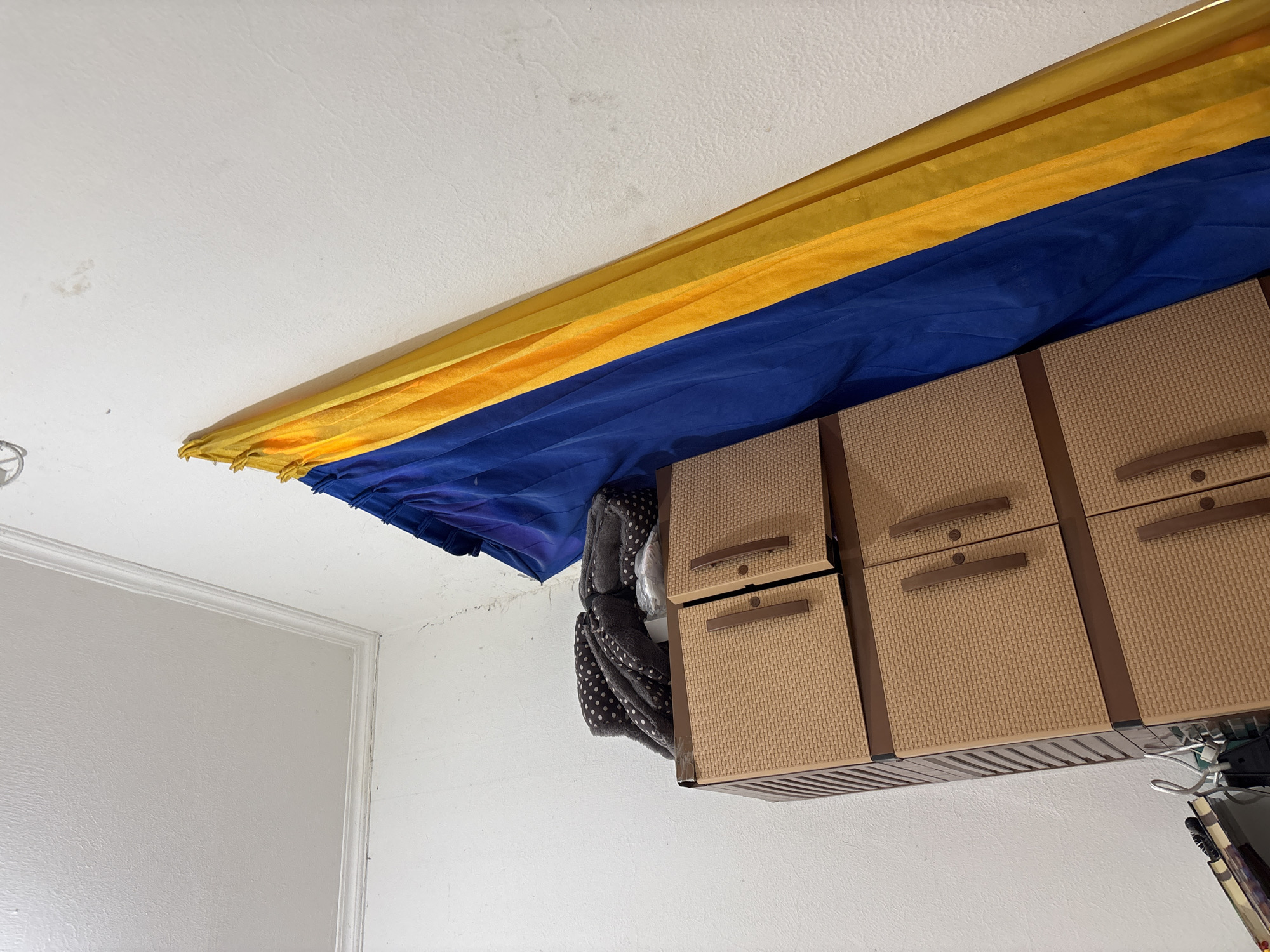 The image depicts an indoor scene, likely a room, in Wanajaya, Indonesia. The walls are white and appear to have some stains or marks, particularly in the upper right quadrant. A white ceiling with molding is visible in the upper left. To the right, a blue and yellow striped curtain hangs prominently, draped against the wall. In the lower portion of the image, a light brown plastic cabinet with woven detailing and dark brown handles is present. On top of the cabinet, there are folded dark fabrics with white polka dots, and some plastic-wrapped items. A book or stack of papers with a white hanger is partially visible in the lower left corner. The scene appears to be a storage area or a corner of a room.