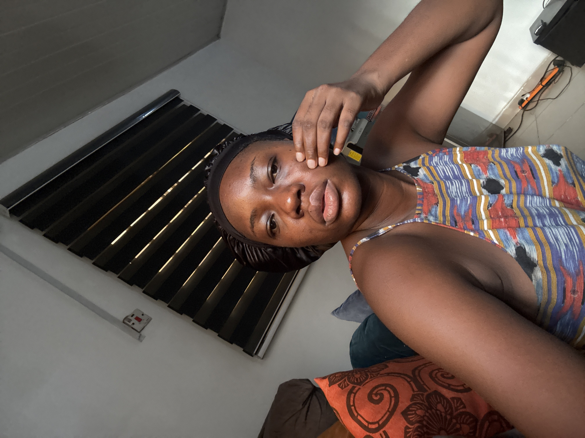 A woman is indoors in Abuja, Nigeria. She wears a patterned tank top and a head wrap. She holds a small object to her face with one hand and her other arm is raised. Blinds are on the wall behind her. Various pillows are behind her on the couch.