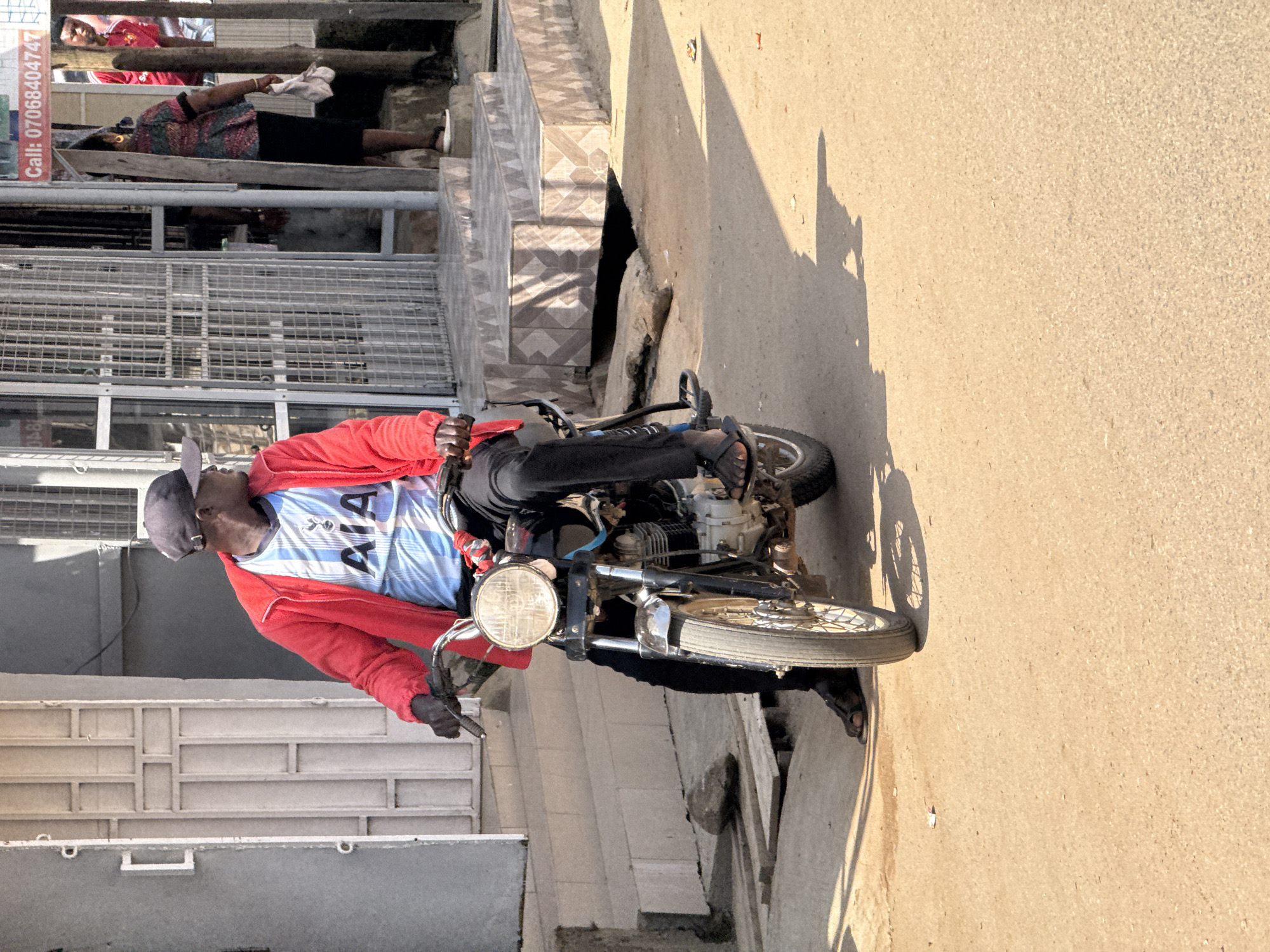 In Abuja, Nigeria, a person is captured on a motorcycle, appearing to be in motion or stopped. The subject is wearing a red jacket, black pants, and a blue and white shirt with "AIA" clearly visible. A dark baseball cap covers their head, and they have sunglasses on. The motorcycle has a prominent round headlight and spoked wheels.
The scene is outdoors, with a paved surface, likely a street or pathway, and a tiled walkway or stairs adjacent to it. The time of day is indicated by the strong shadows cast by the motorcycle and a person's silhouette on the ground, suggesting it is either morning or late afternoon with the sun low in the sky. The sunlight is bright, illuminating the scene and casting distinct shadows.
In the background, there are glimpses of buildings or structures with metallic fencing and windows. A red and white sign with vertical text, though partially obscured, is visible on the left side of the frame. The overall atmosphere appears to be a typical street scene in a populated urban environment. There are no overt signs of distress or celebration, suggesting a commonplace event.