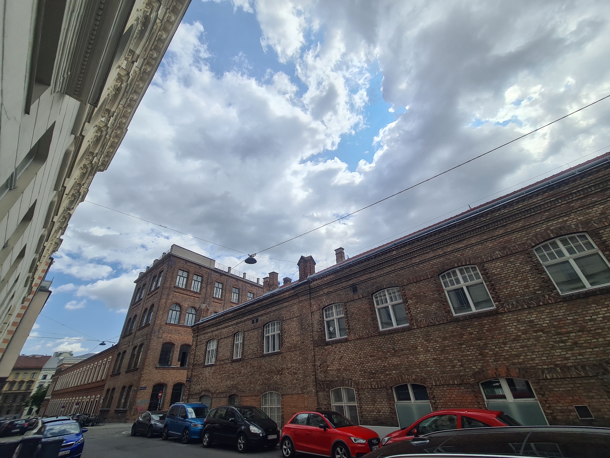 The image depicts a street scene in Vienna, Austria, featuring an angled view of historic brick buildings and parked cars under a cloudy sky. On the left, a light-colored building with decorative architectural elements lines the street. To the right, a long, multi-story brick building with numerous windows, some arched and some rectangular, dominates the midground and background. Several cars are parked along the street, including a blue hatchback, a black hatchback, a red hatchback, and a dark-colored sedan. The overall atmosphere suggests an urban environment with a sense of historical depth. The buildings' architecture and the street appear to be in a relatively well-maintained condition.